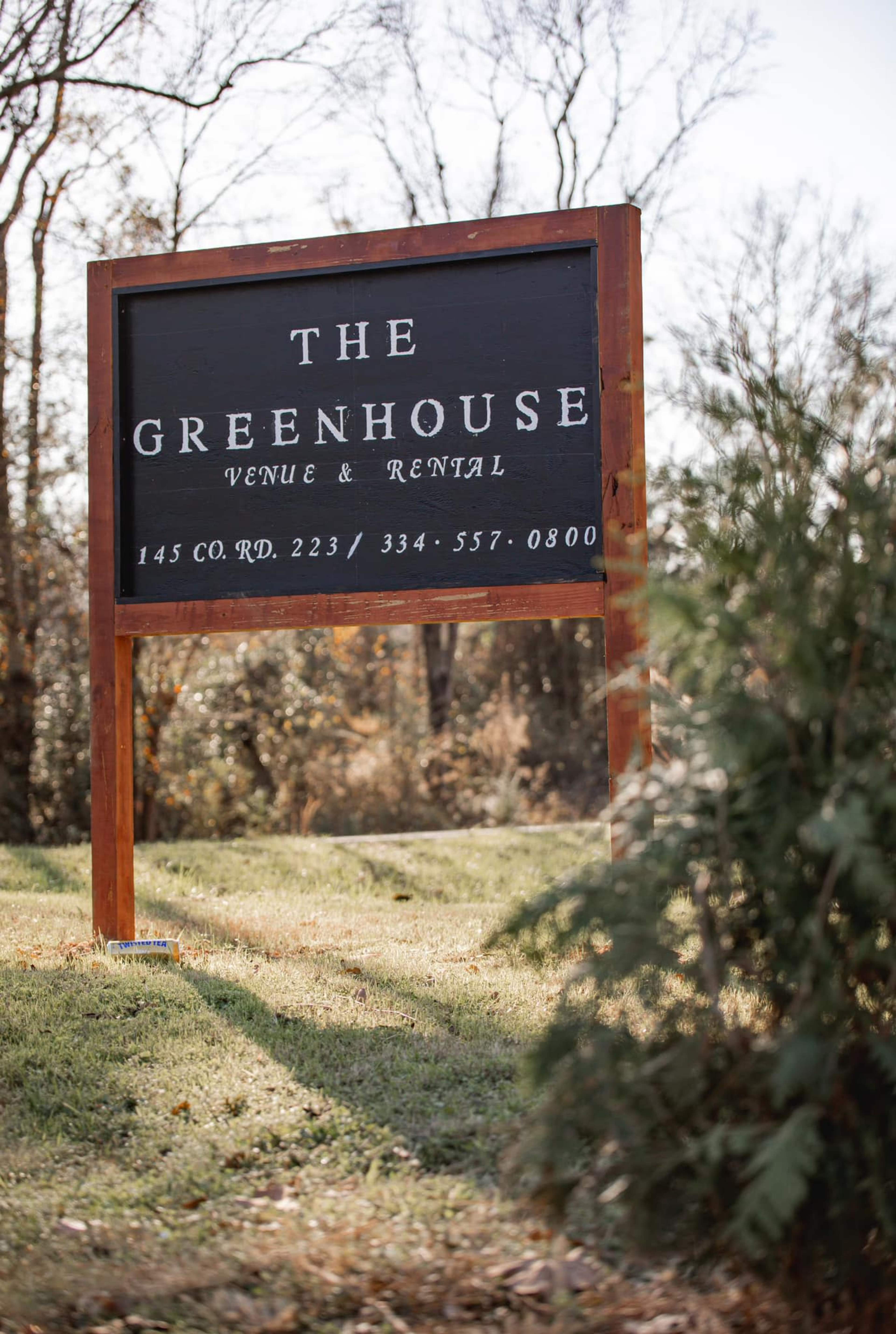 A wooden sign reads "The Greenhouse Venue & Rental" with contact information, set in a grassy area among trees.