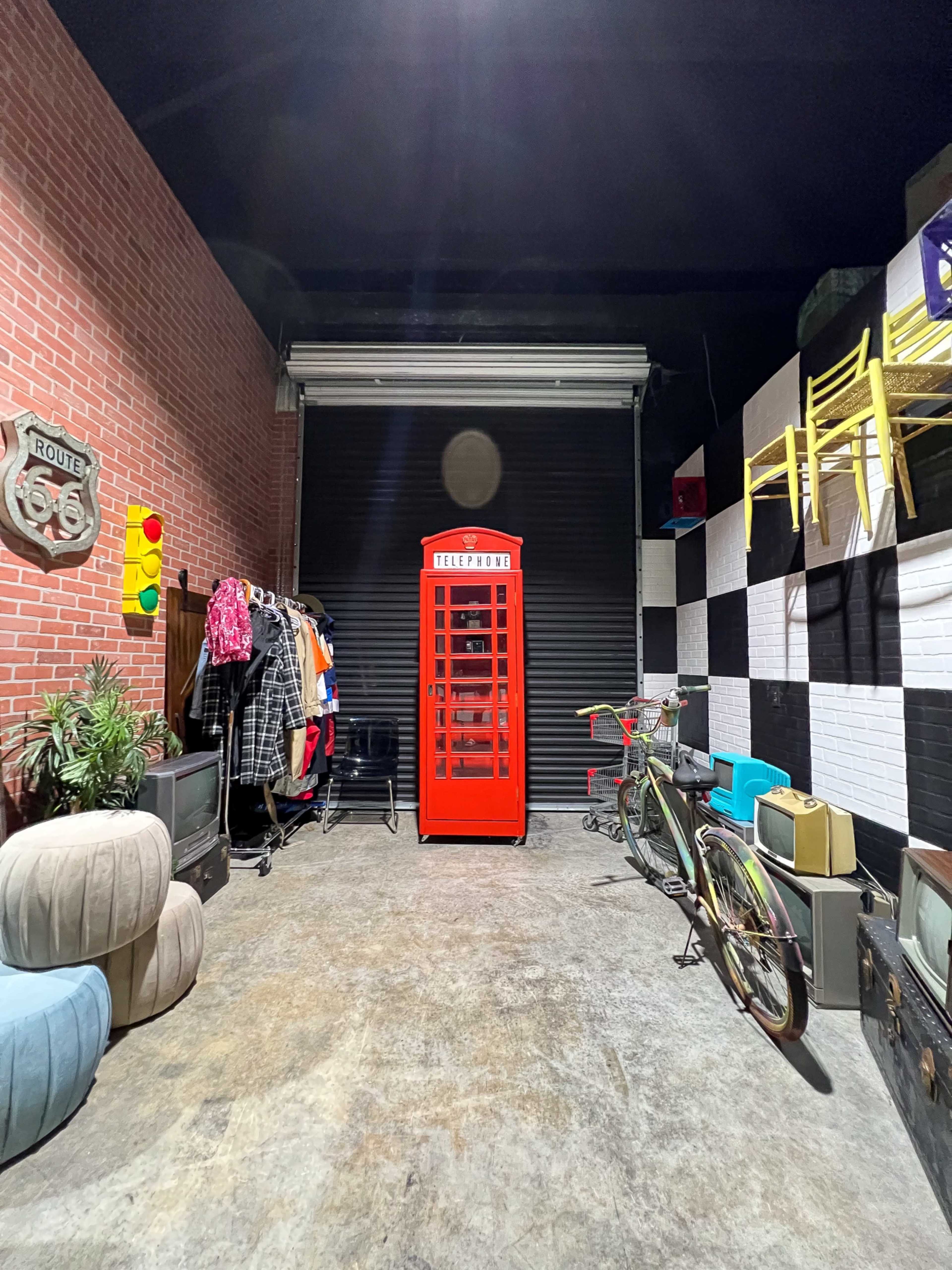 The image shows a vintage red telephone booth in a garage filled with various clothing items, bicycles, and furniture against a backdrop of brick and checkered walls.