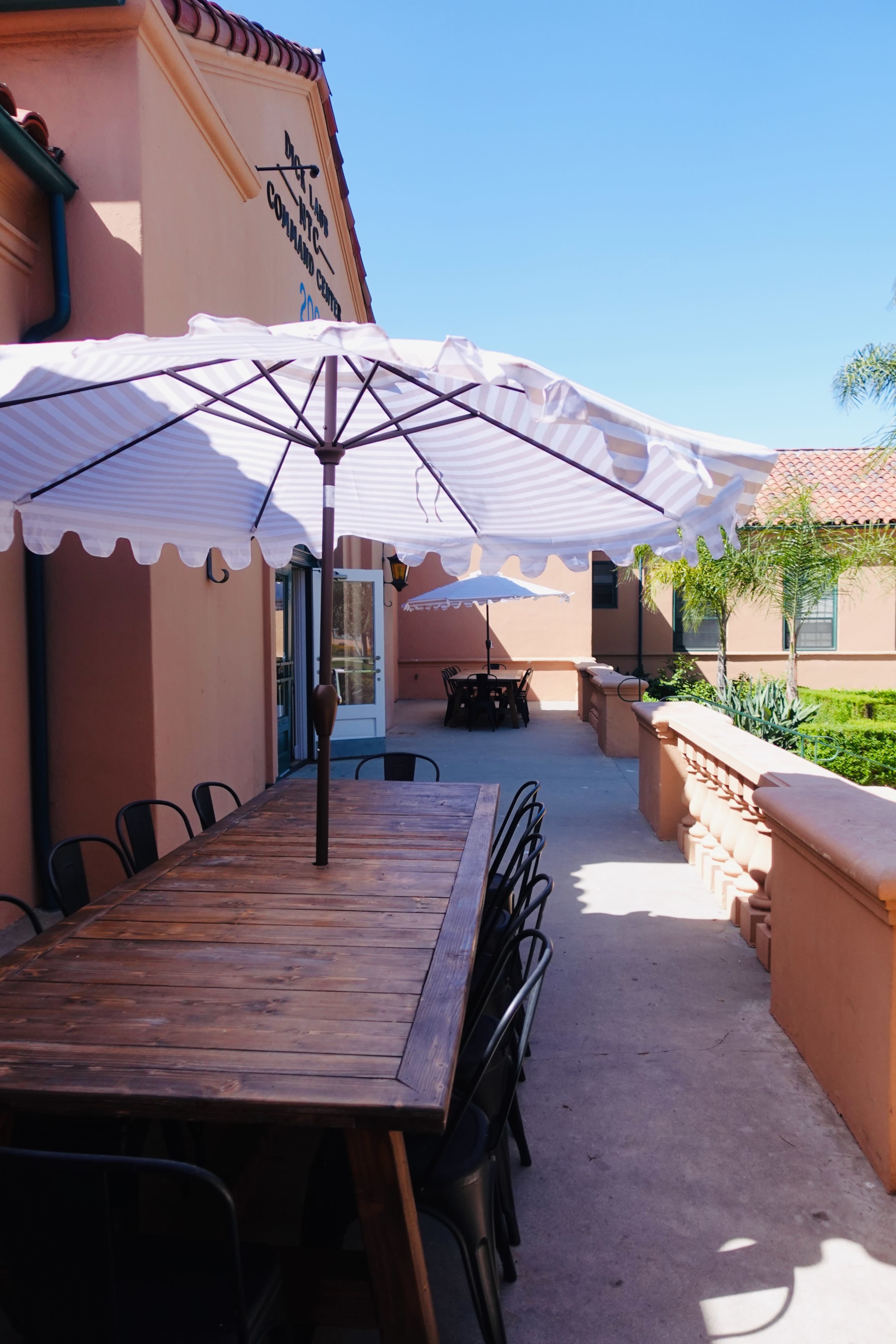 A wooden table with black chairs and large umbrellas is situated along a pathway next to a building with a stucco exterior.