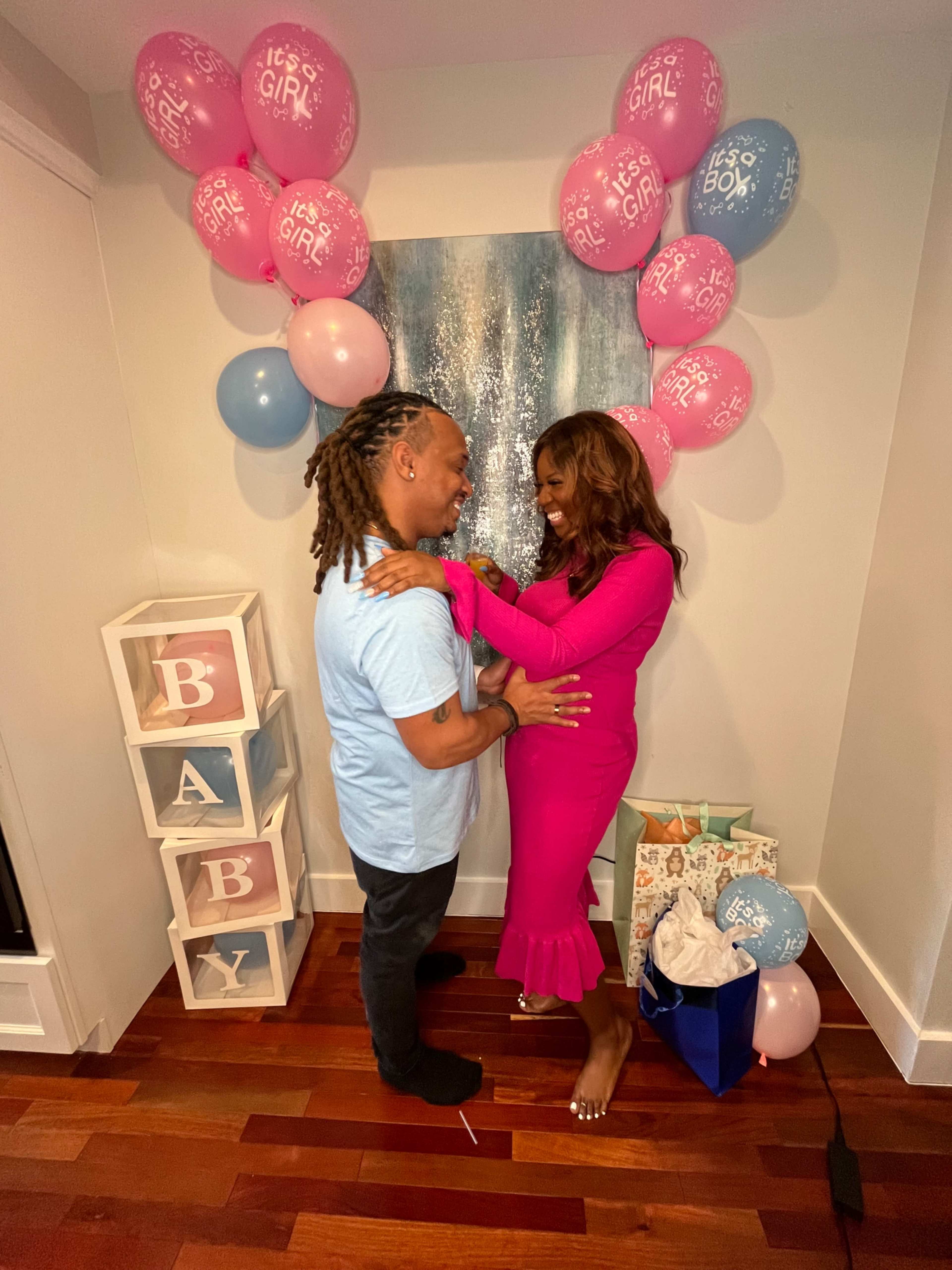 A couple stands together in a decorated room with pink and blue balloons and a display that reads "BABY."