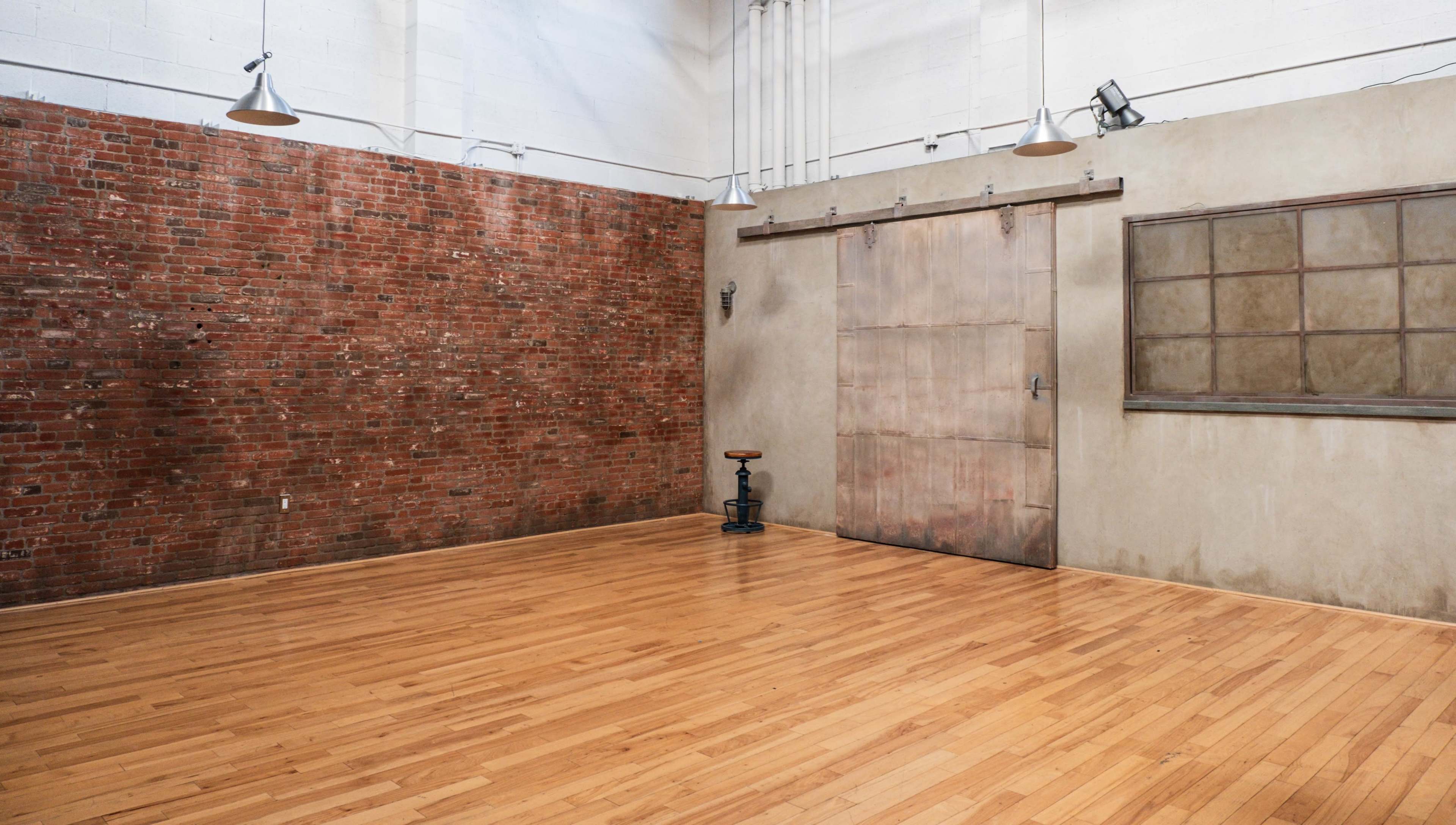 A spacious, empty room with a wooden floor, a brick wall, and a metal sliding door next to a window.