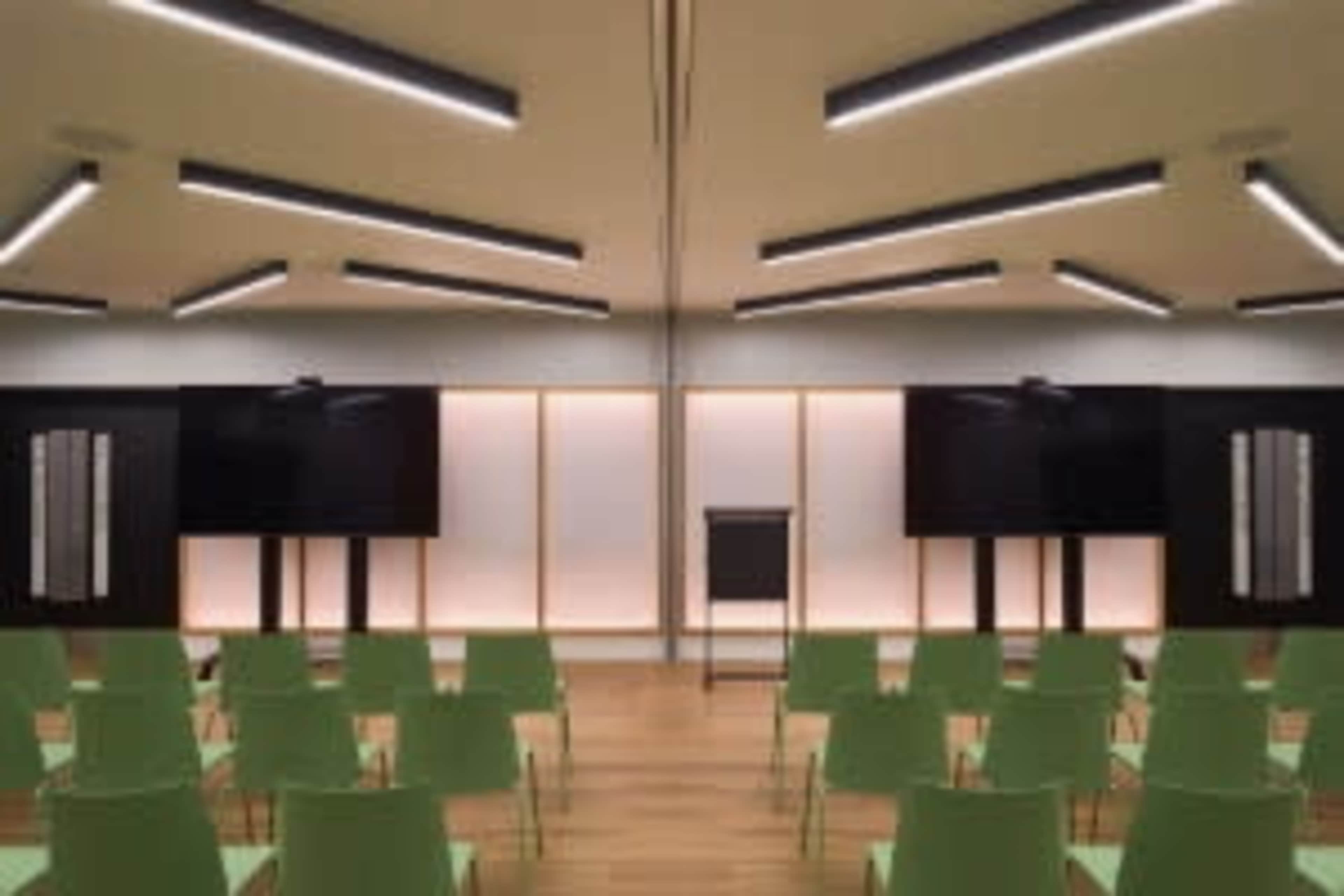 The image shows a modern conference room set up with green chairs arranged in rows facing two large screens and a podium.