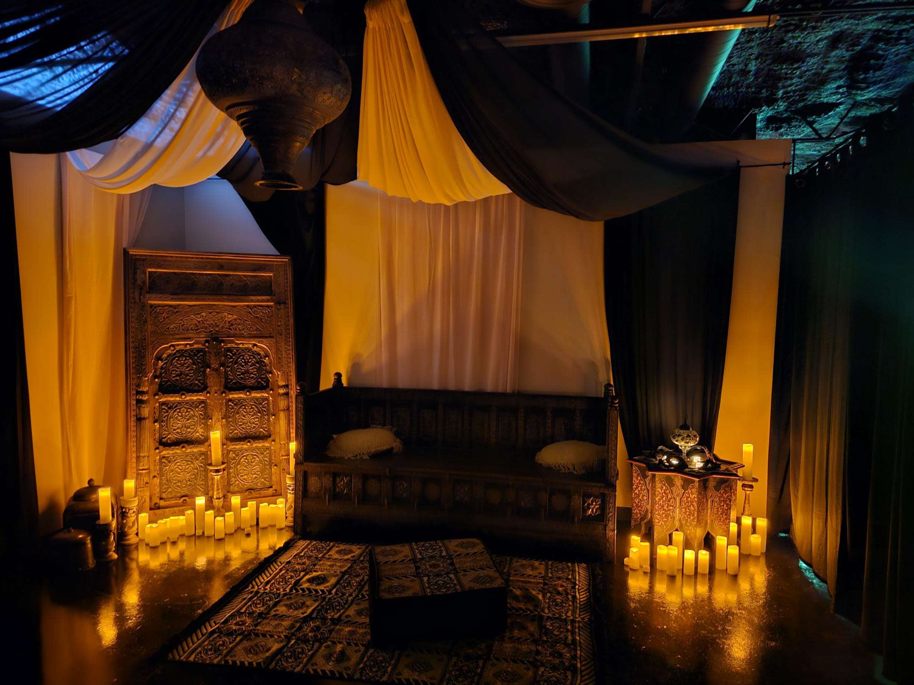 A softly lit interior with a decorative wooden couch, surrounded by candles and draped fabric in various colors.