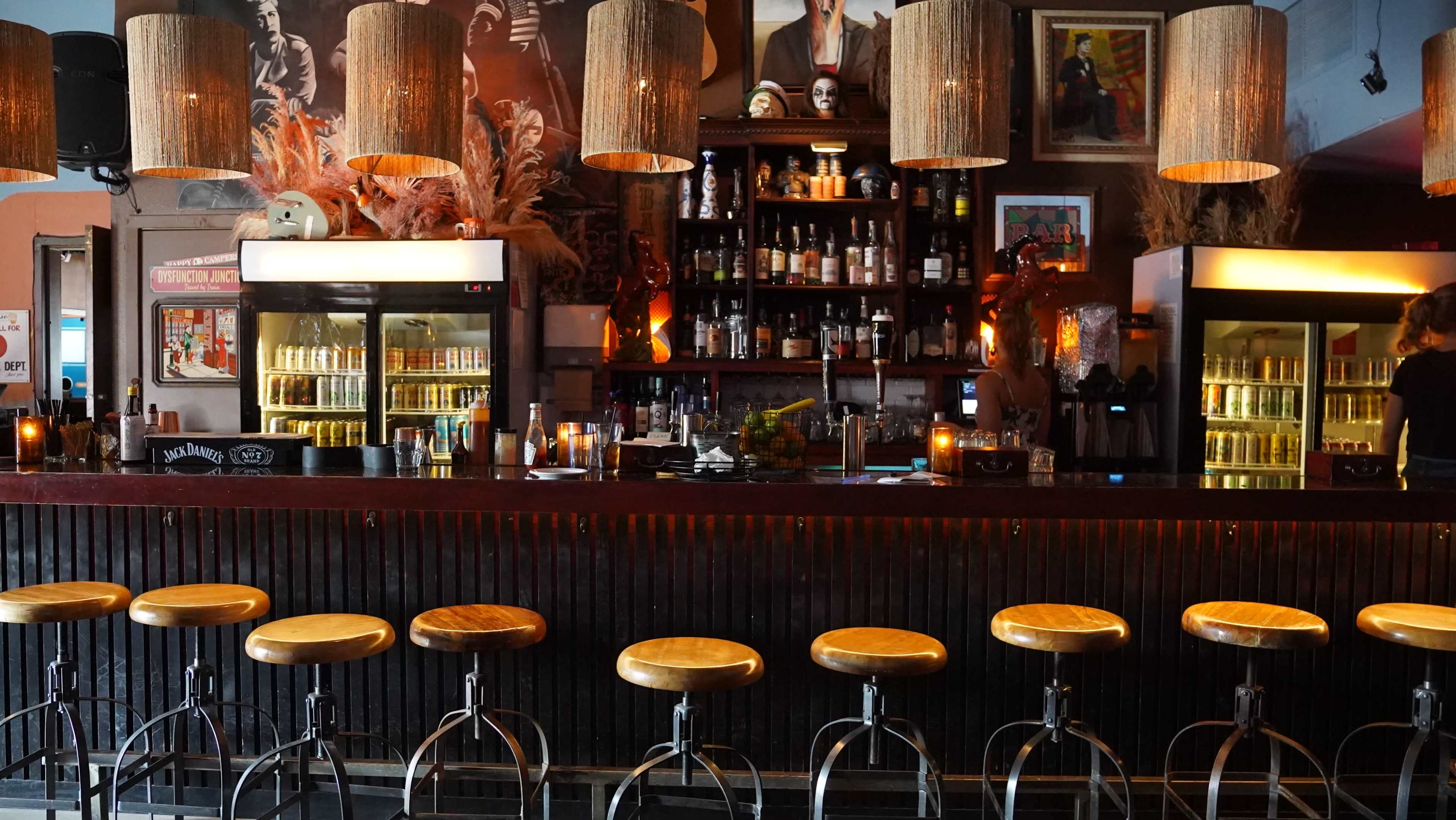 The image shows a bar with wooden stools lined up in front of a dark wooden counter, illuminated by pendant lights and featuring shelves filled with various bottles behind it.