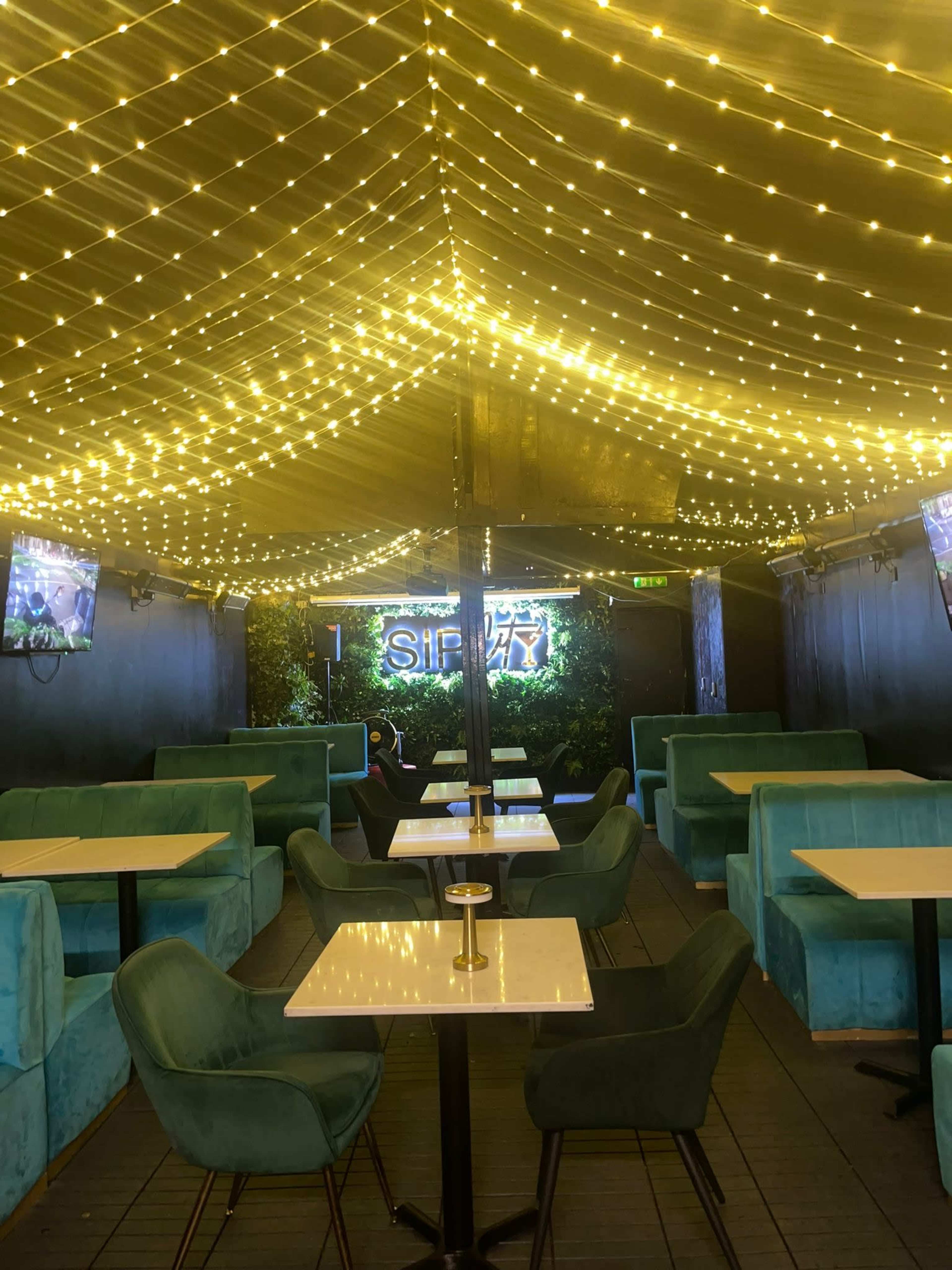 The image shows a dimly lit indoor venue with teal seating, string lights overhead, and a neon sign that reads "SIP".