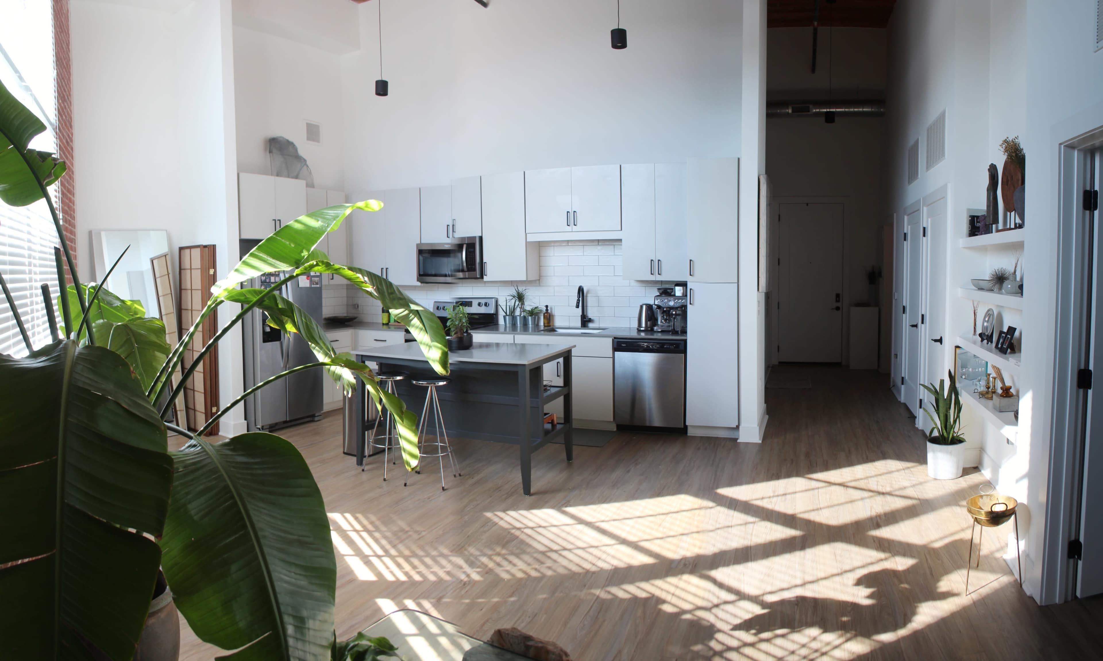 A bright, open kitchen features white cabinets, a gray island, and large windows casting shadows on the wooden floor.