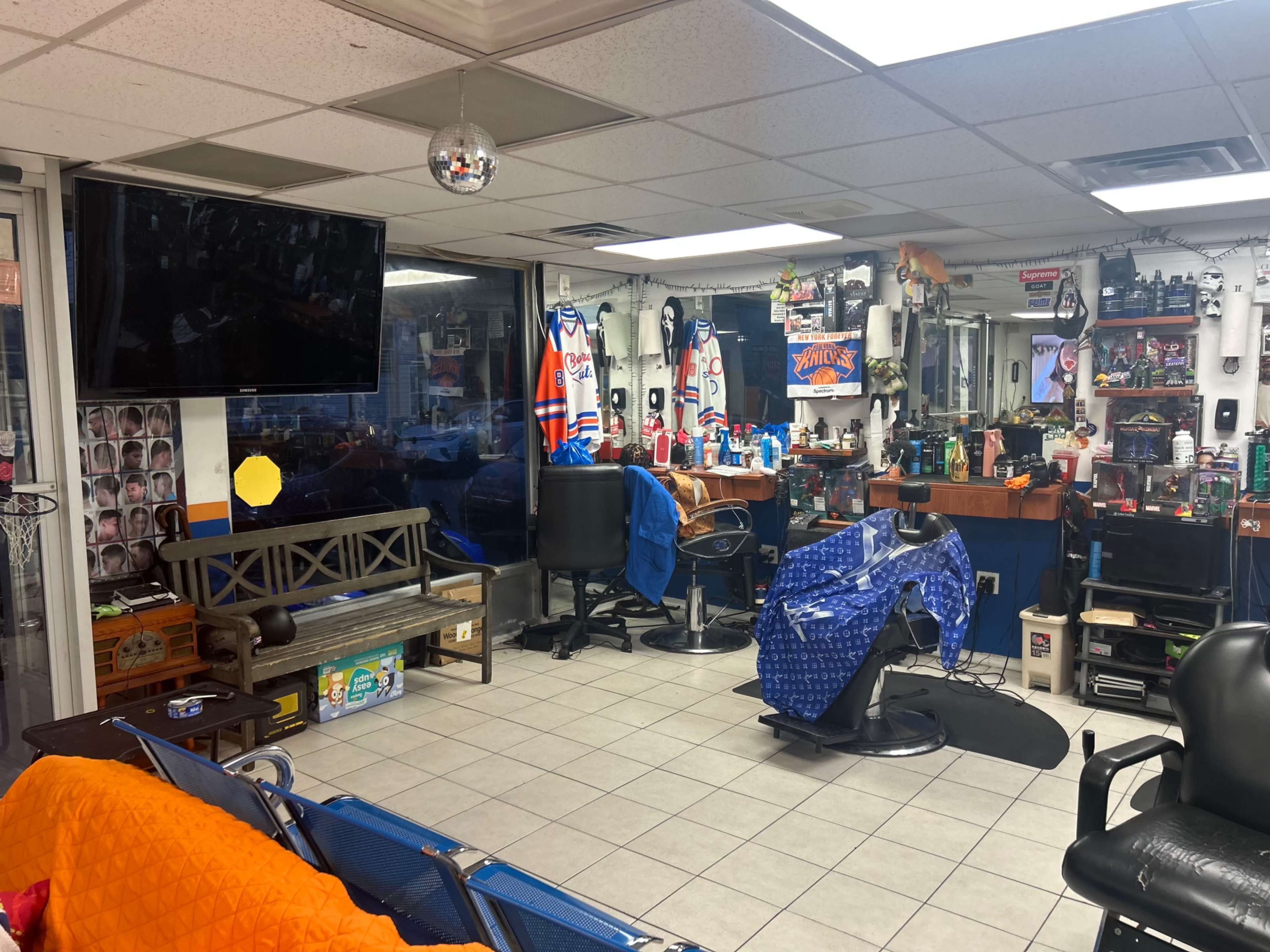 The interior of a barbershop features a waiting area with chairs, a large flat-screen TV on the wall, and various grooming supplies organized on counters.