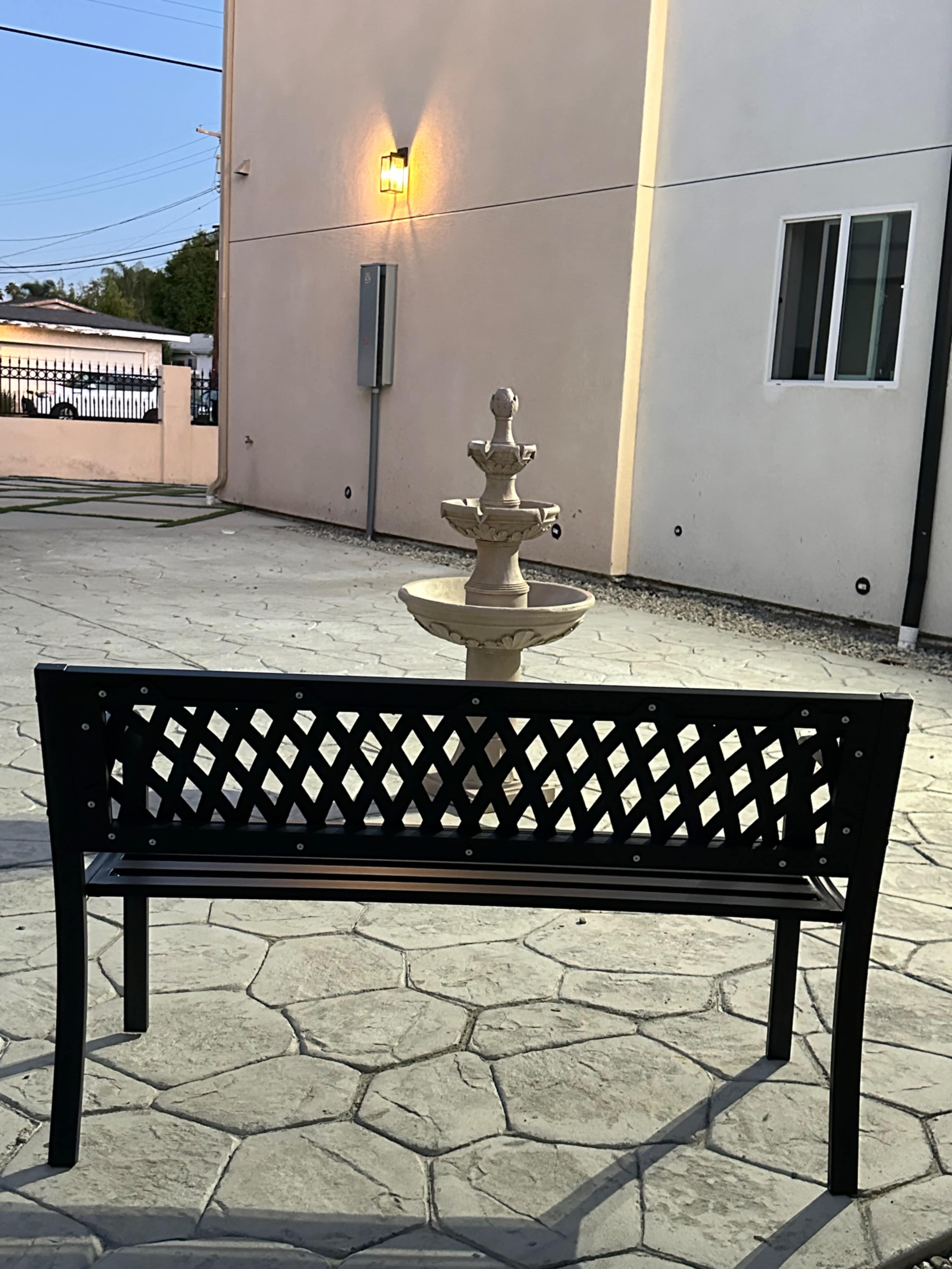 The image shows a black metal bench facing a stone fountain in a patio area, with a building wall in the background.