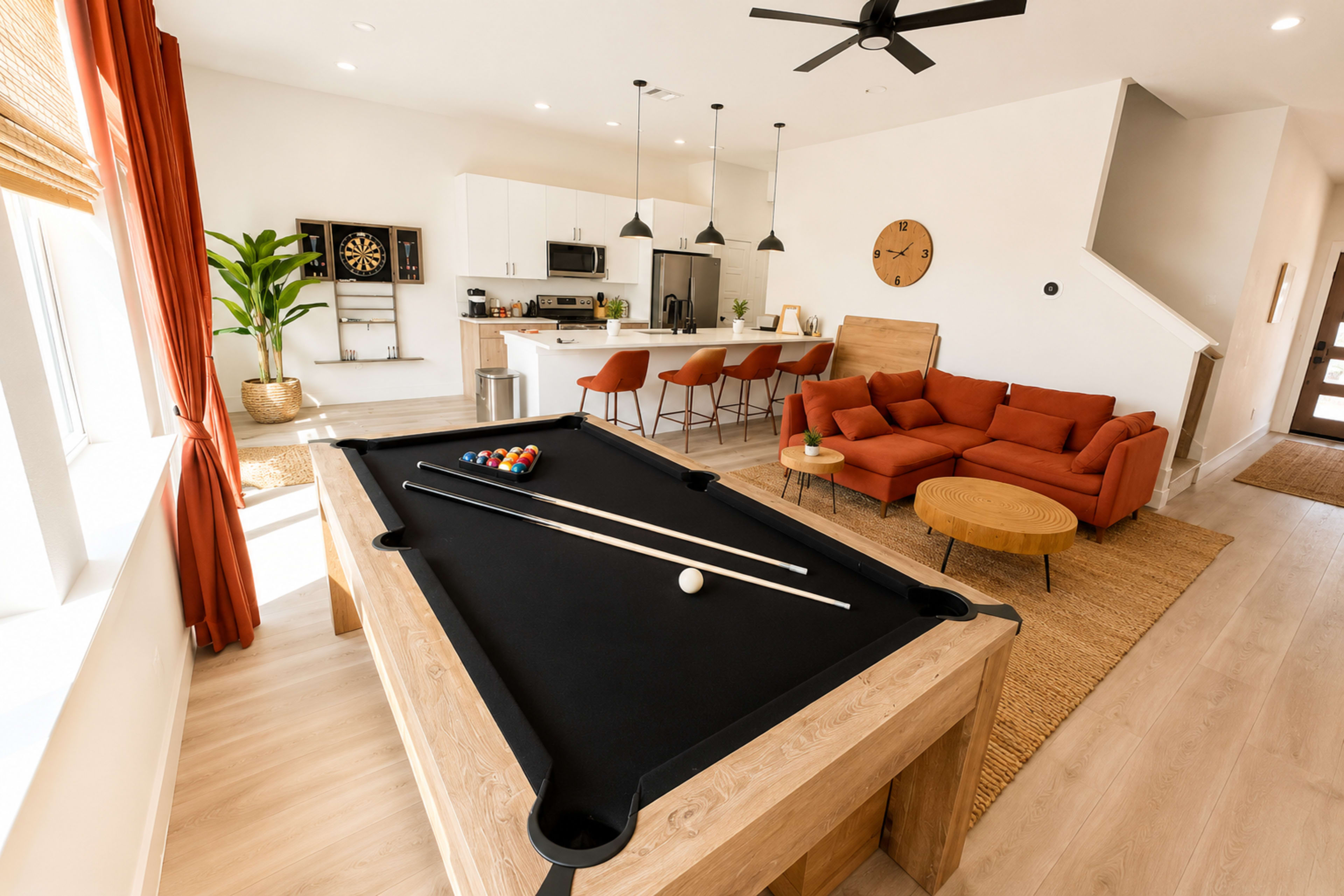 A spacious room features a pool table in the foreground, with a modern kitchen and living area visible in the background.