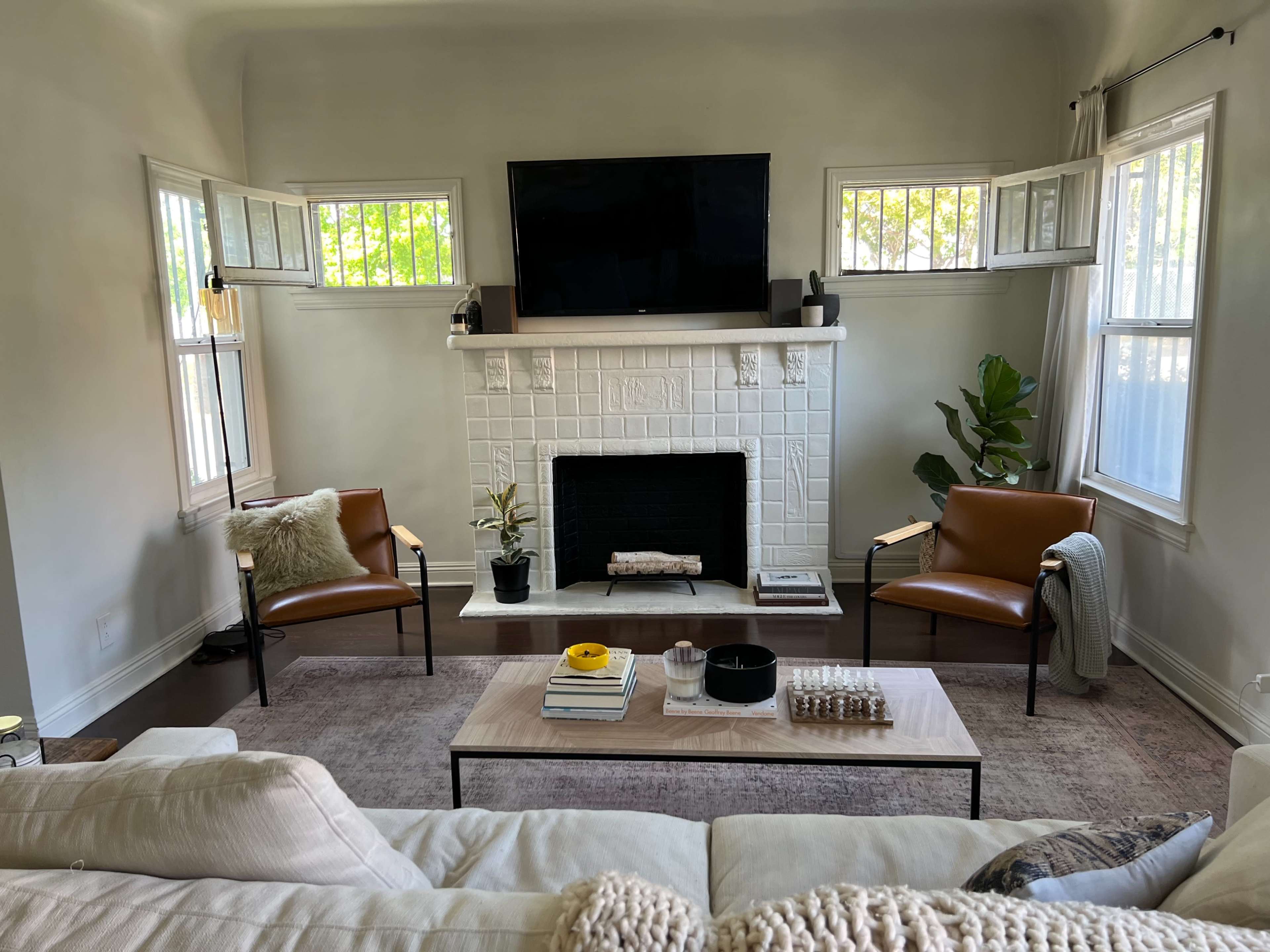 The image shows a bright living room with a white brick fireplace, two brown armchairs, a coffee table, and a television mounted above the fireplace.