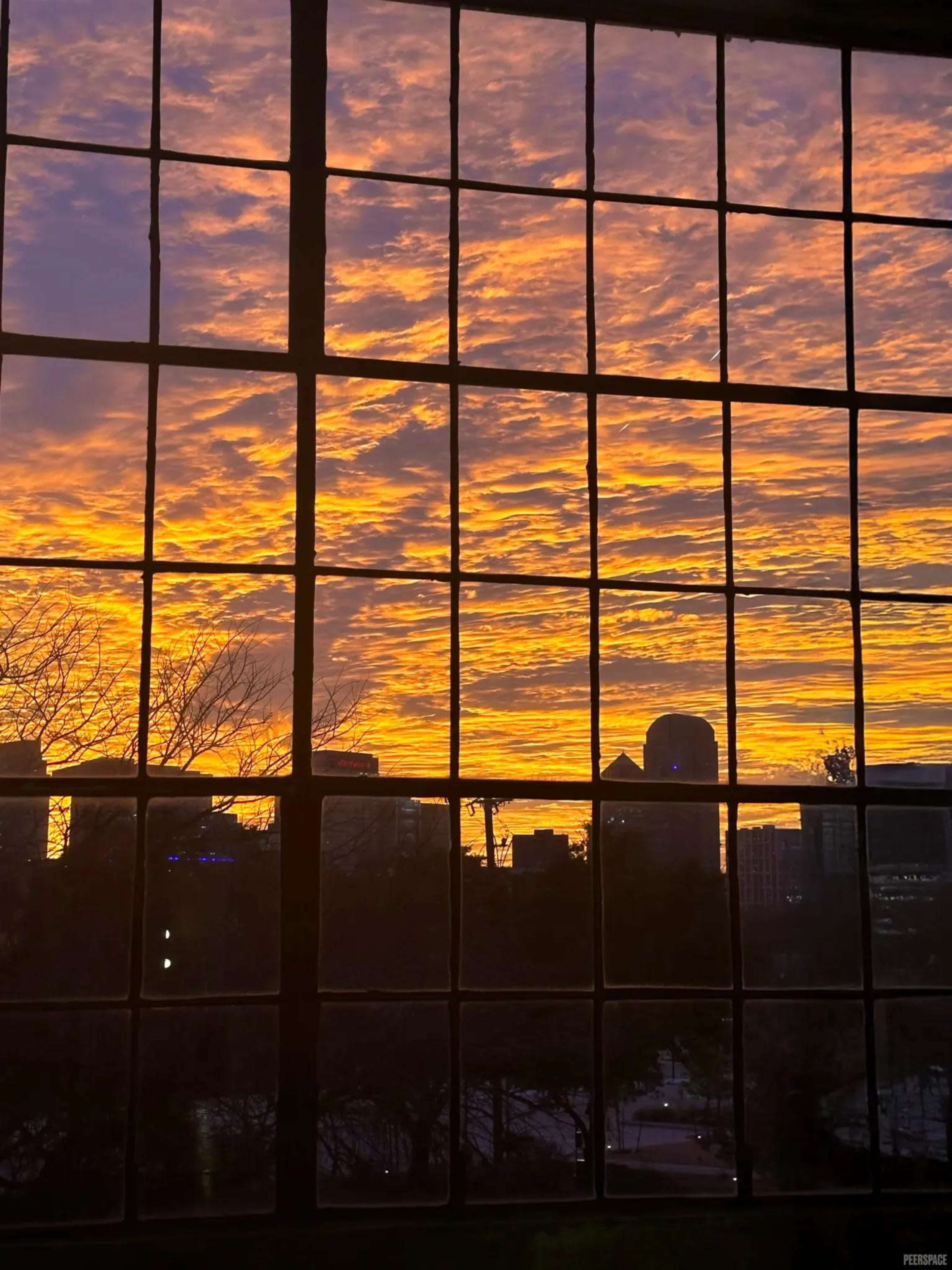 The image shows a stunning sunset with vibrant orange and purple clouds silhouetted against a city skyline viewed through a large window with grid panes.