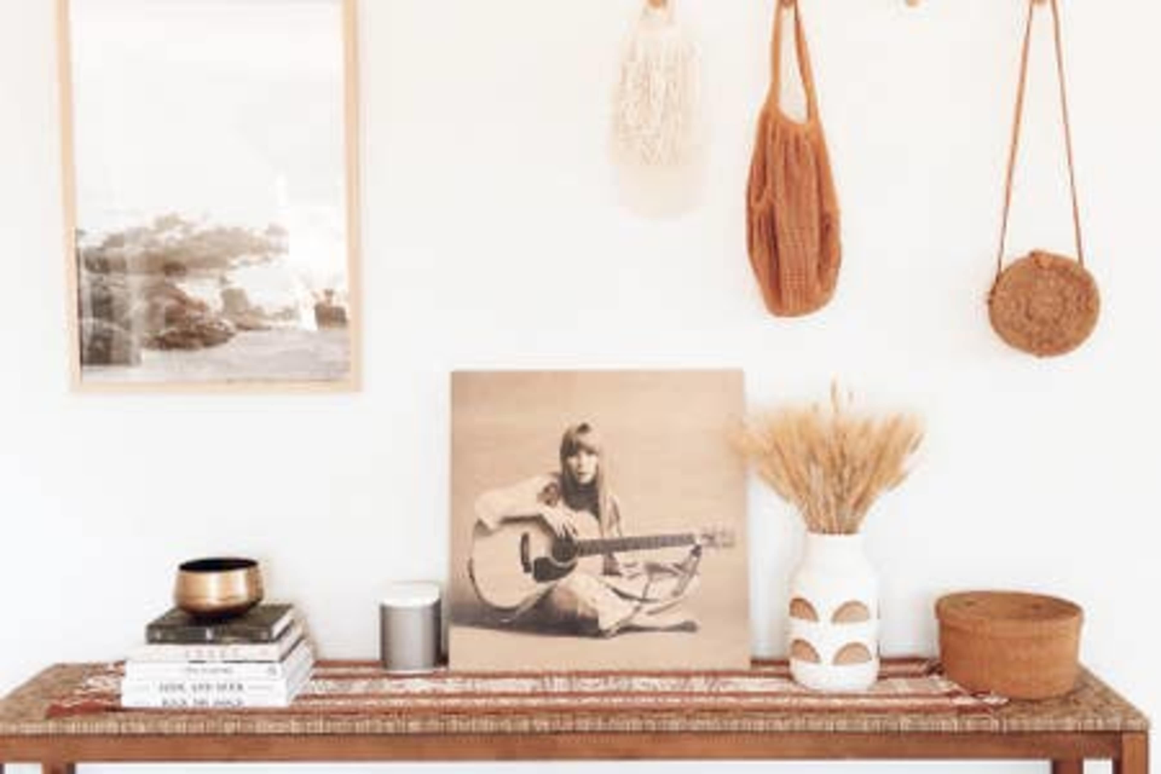 A wooden table displays a framed photograph, a vintage album cover of a woman playing guitar, various decorative items, and a vase with dried flowers against a white wall.