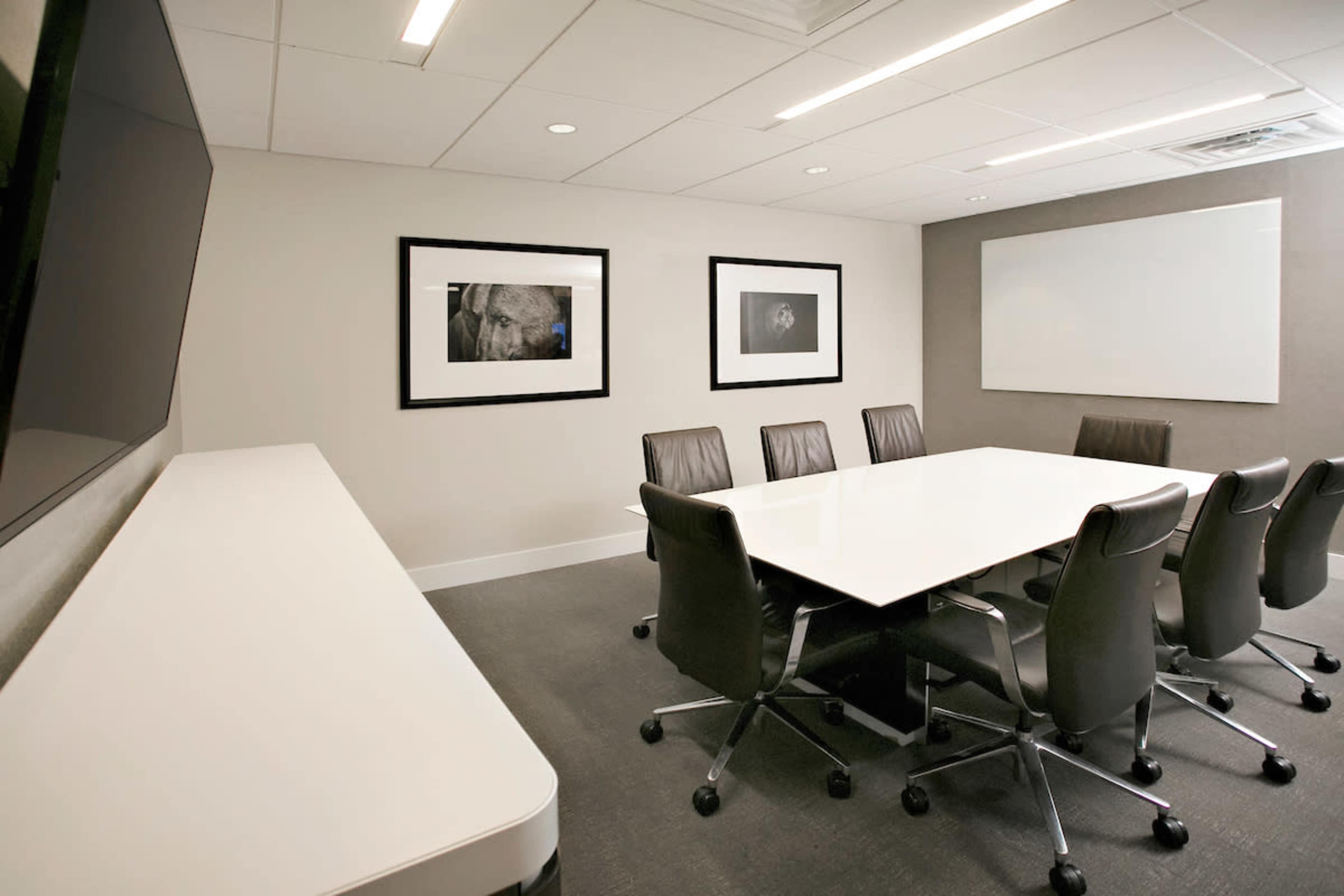 The image shows a modern conference room with a white table surrounded by seven black office chairs and framed photographs on the walls.