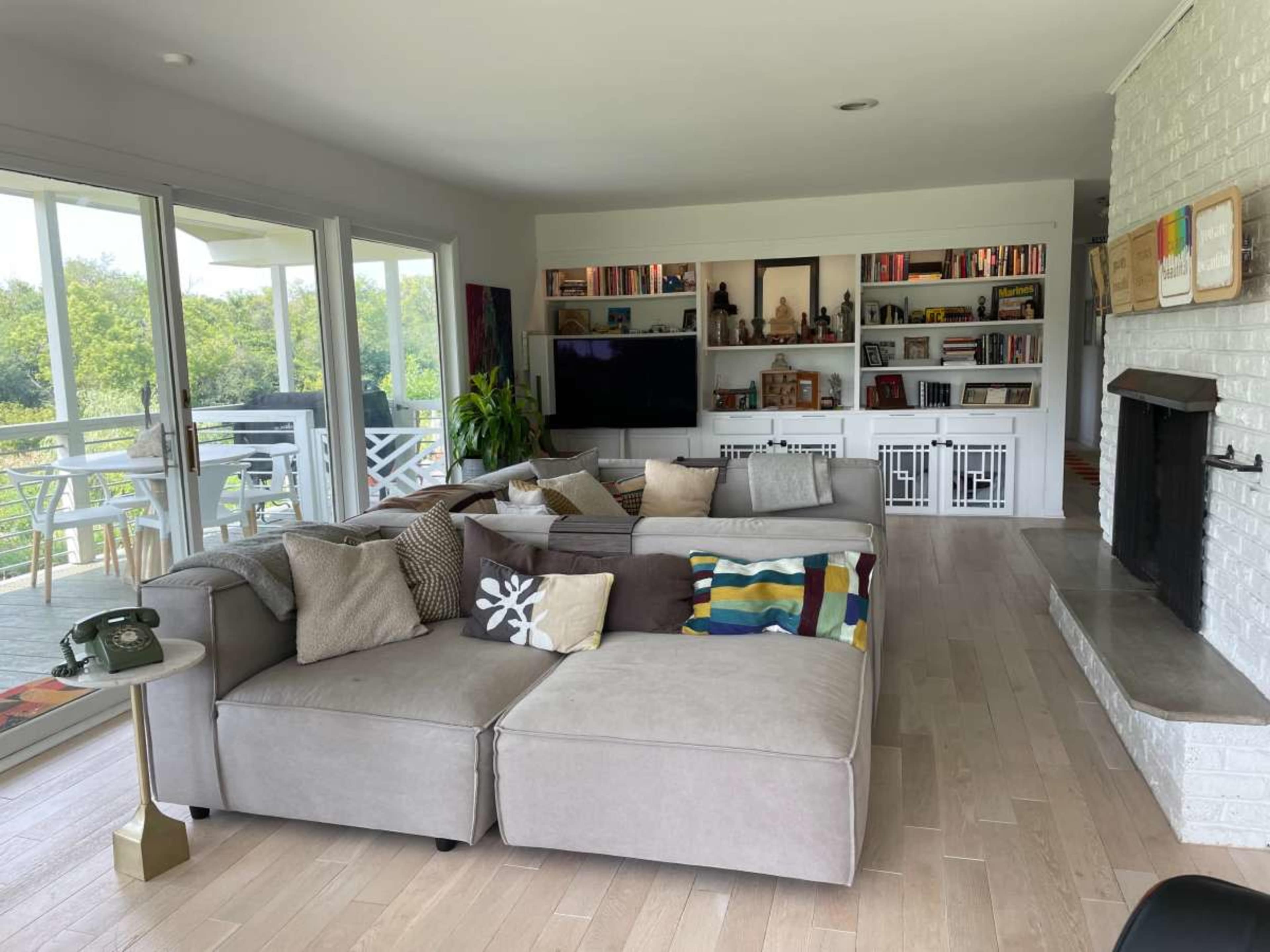 A spacious living room with a large gray sectional sofa, a coffee table, a shelf filled with books, and a glass door leading to a balcony.