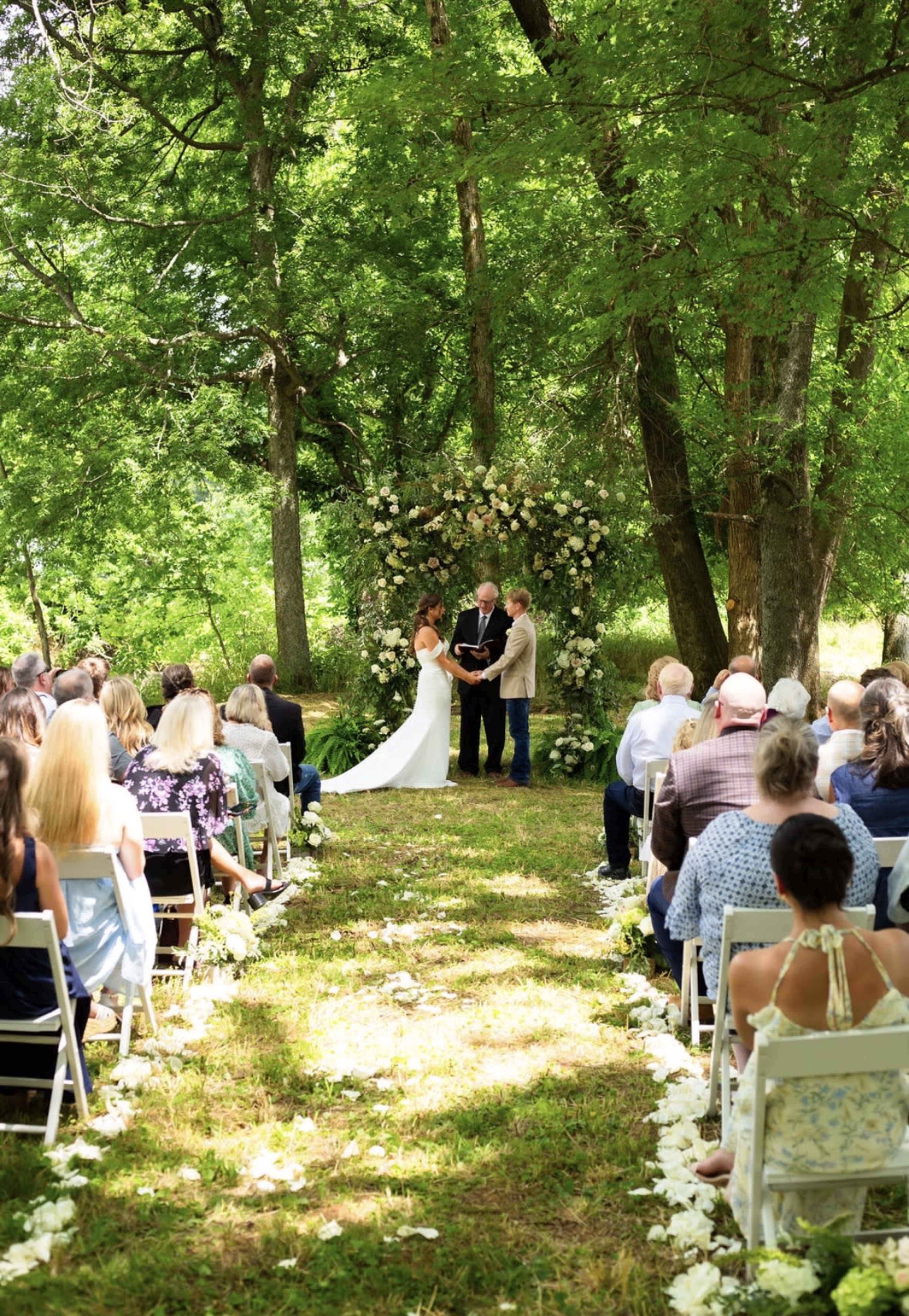 A couple stands under a floral arch while exchanging vows during an outdoor wedding ceremony in a lush green setting.