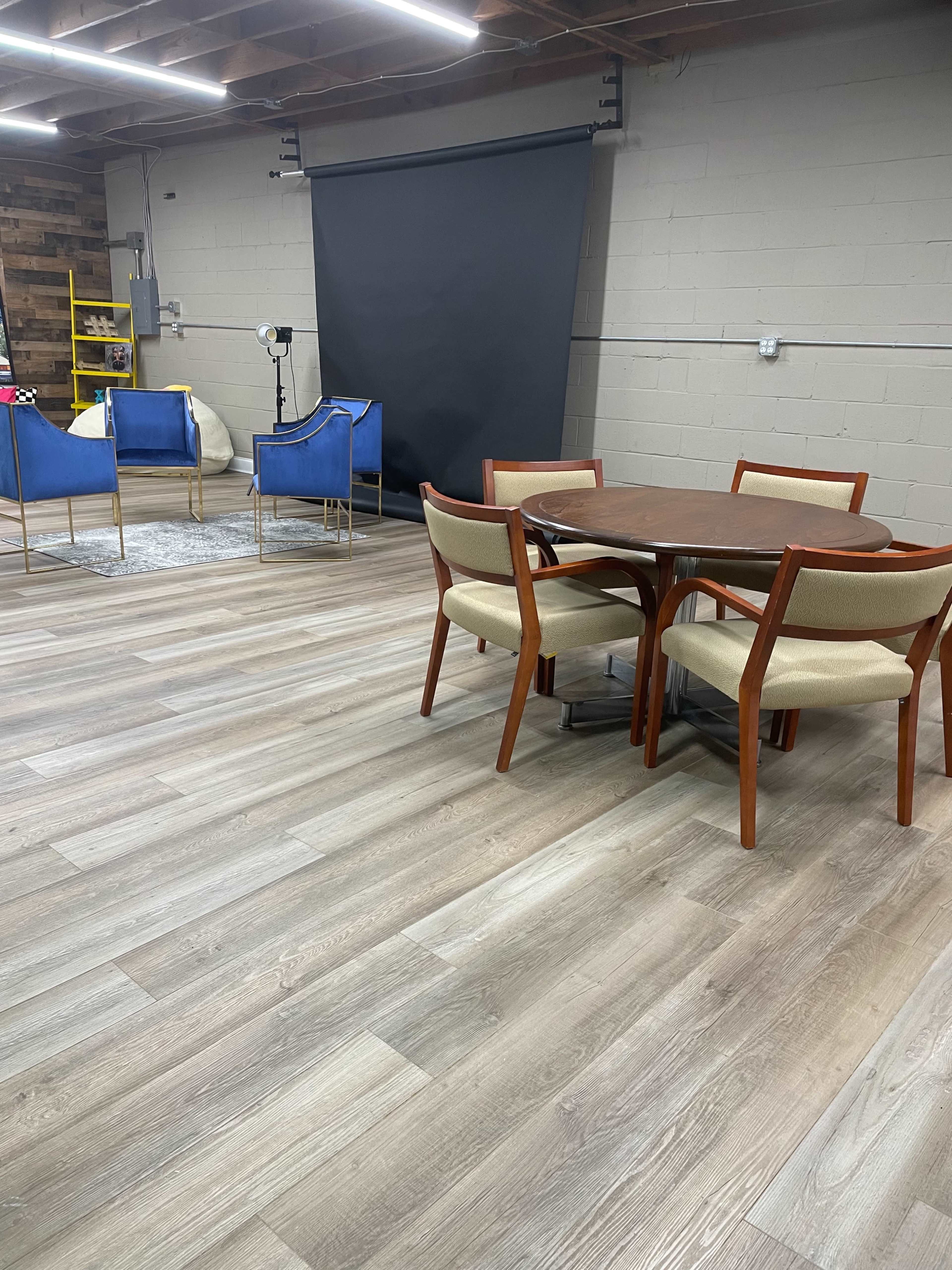 A spacious room with gray wood flooring, featuring a round wooden dining table surrounded by four upholstered chairs, and two blue armchairs in the background near a black backdrop.
