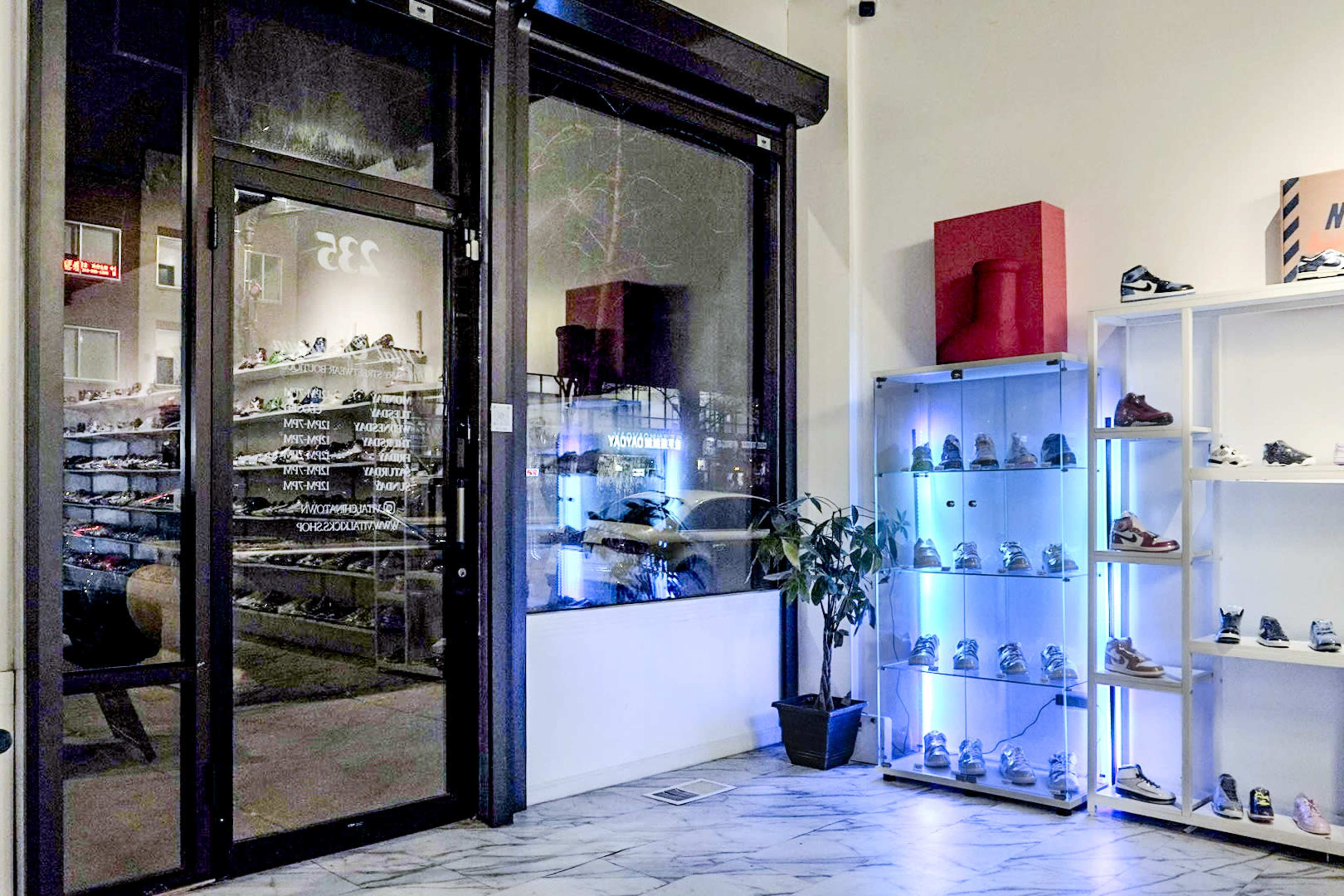 The interior of a sneaker store featuring a glass door entrance, a display of shoes on shelves, and a plant in the corner.
