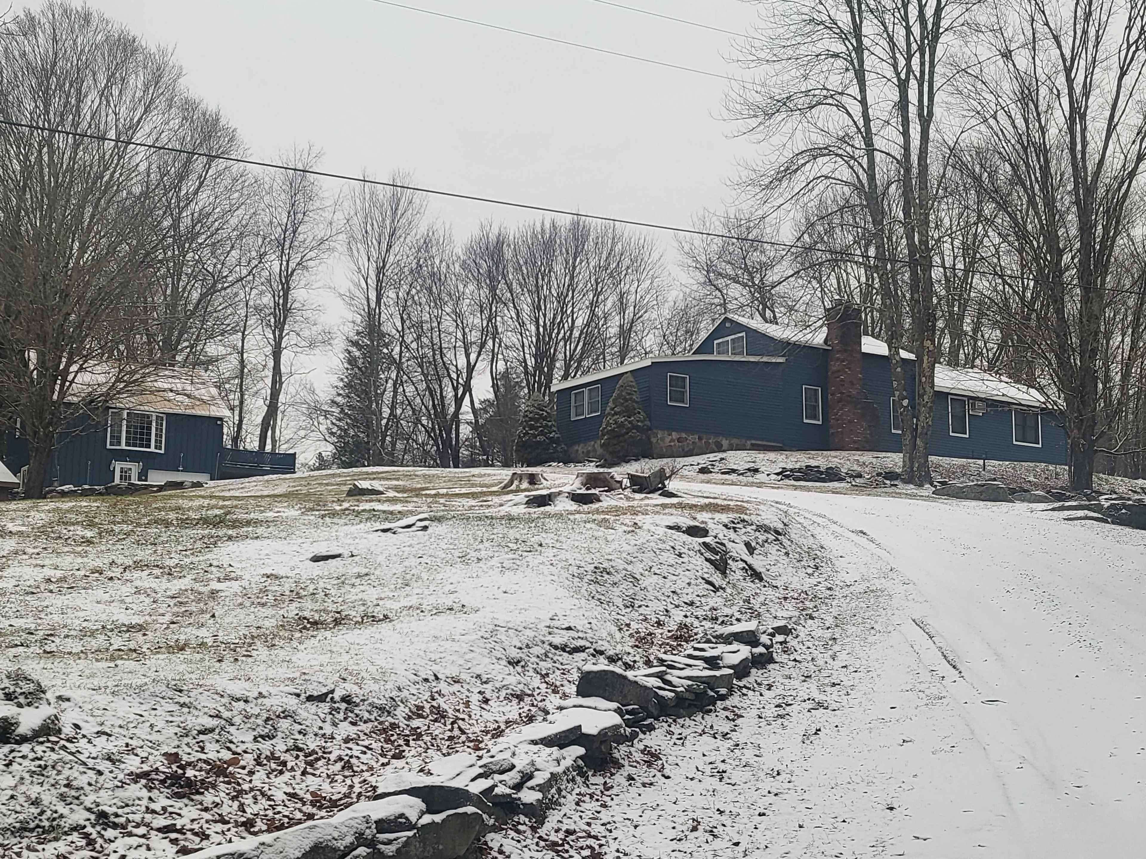 A blue house with a stone chimney is situated on a snow-covered hillside surrounded by bare trees.