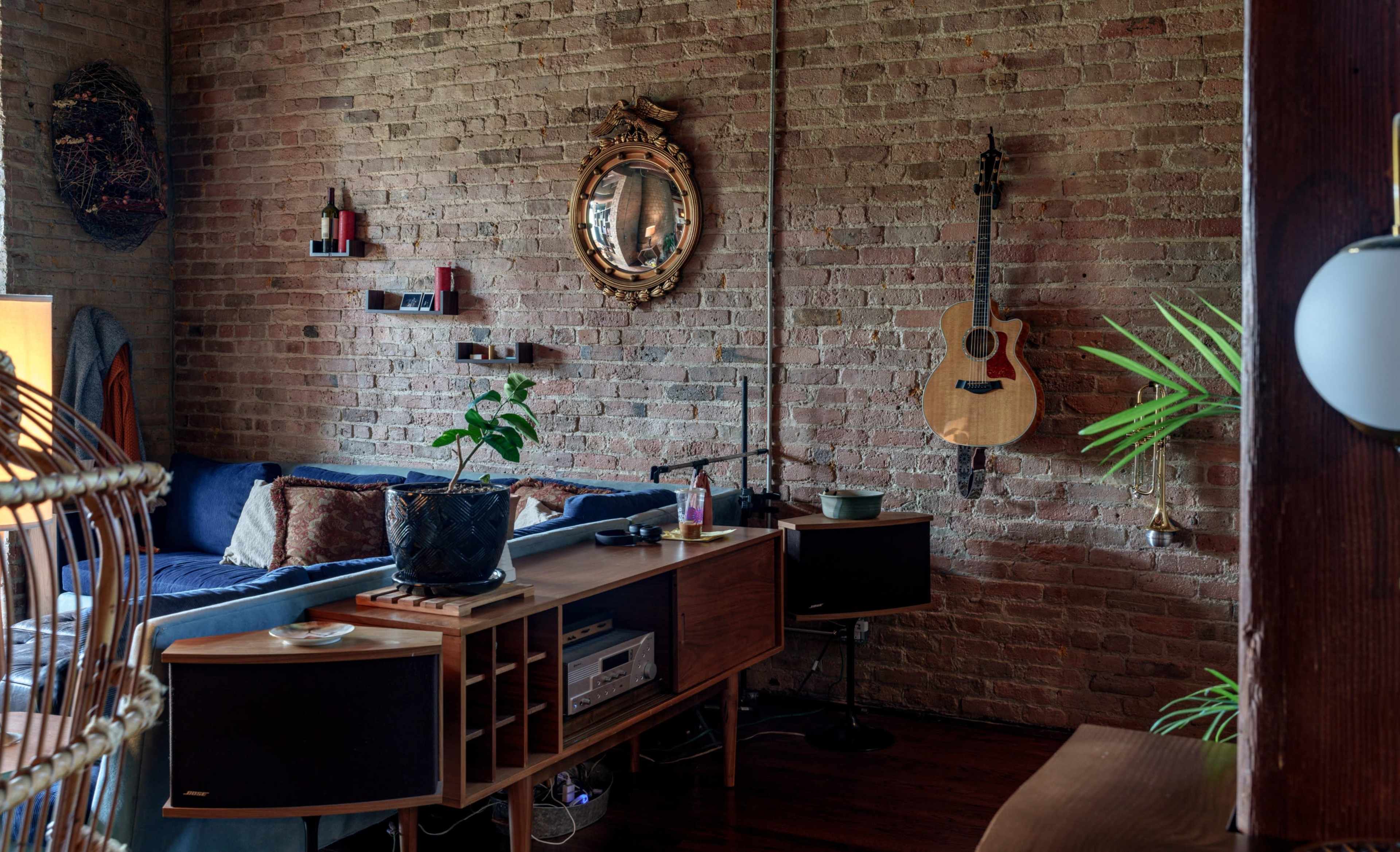 The image shows a cozy living room featuring a brick wall, a blue sofa, a wooden console table, and an acoustic guitar hanging on the wall.