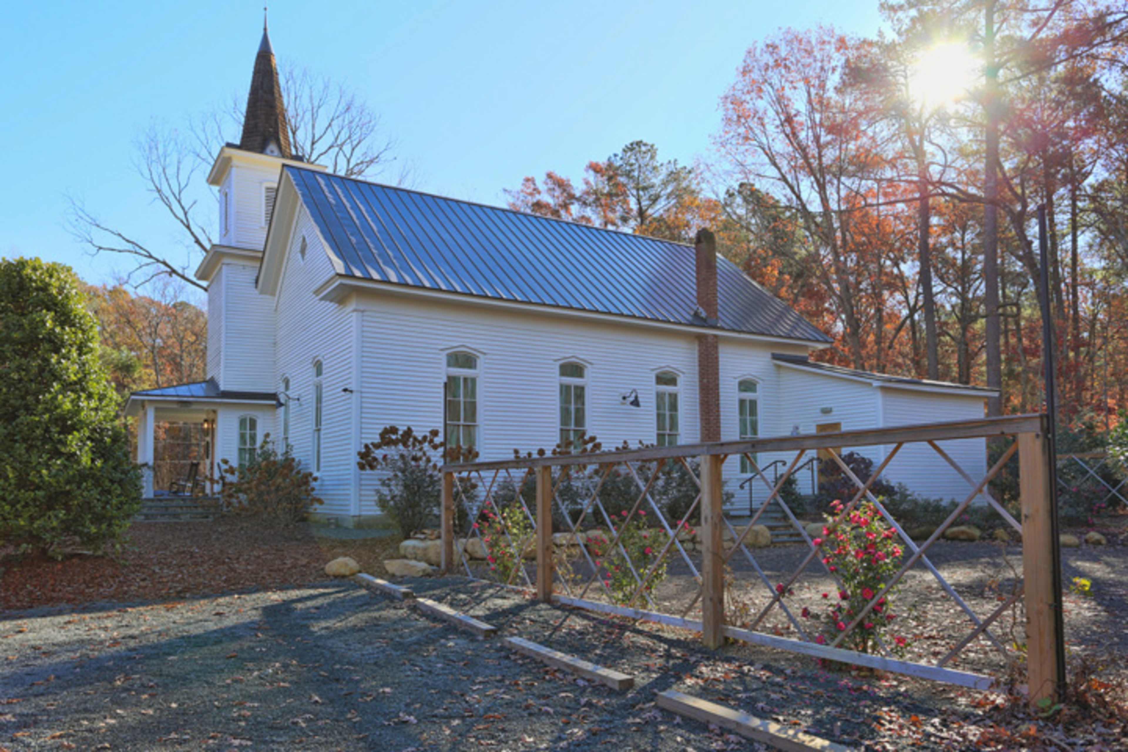 A white church with a steeple is surrounded by trees displaying autumn foliage and features a gravel pathway with a wooden fence in the foreground.