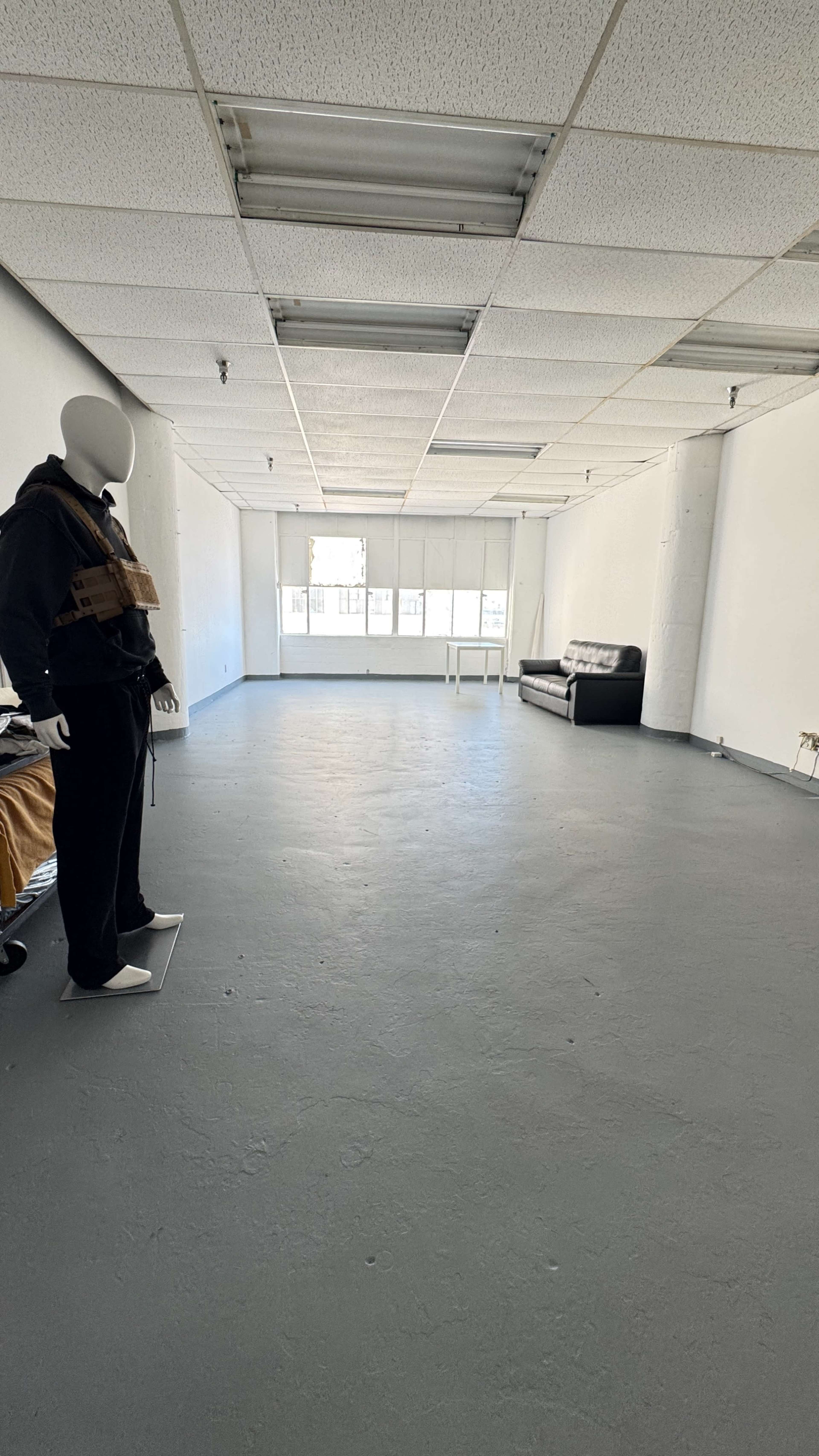The image shows an empty room with gray flooring, a mannequin dressed in dark clothing, and a black sofa against one wall.