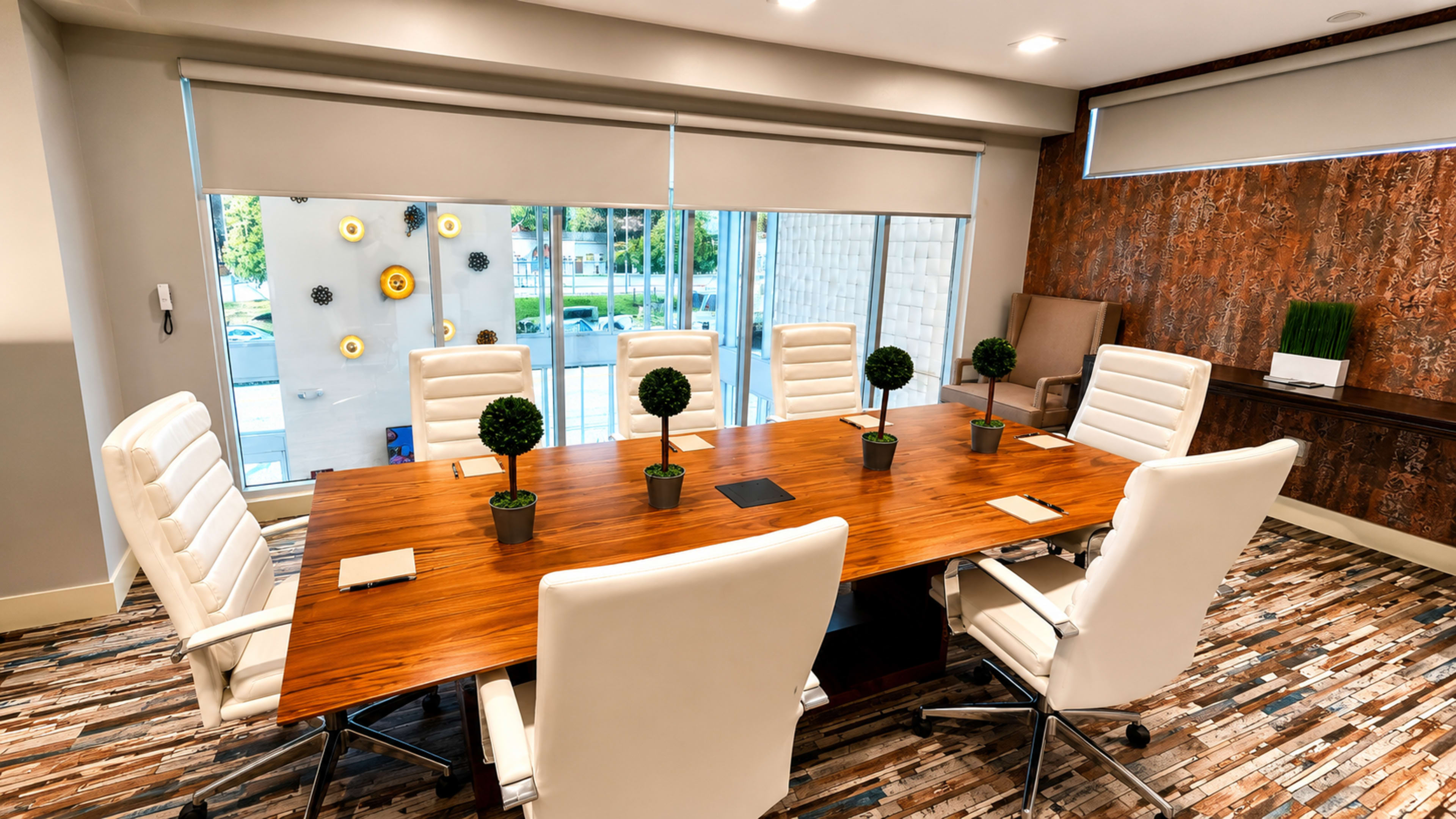 A modern conference room features a large wooden table surrounded by white executive chairs, with decorative plants in small pots on the table and large windows offering natural light.