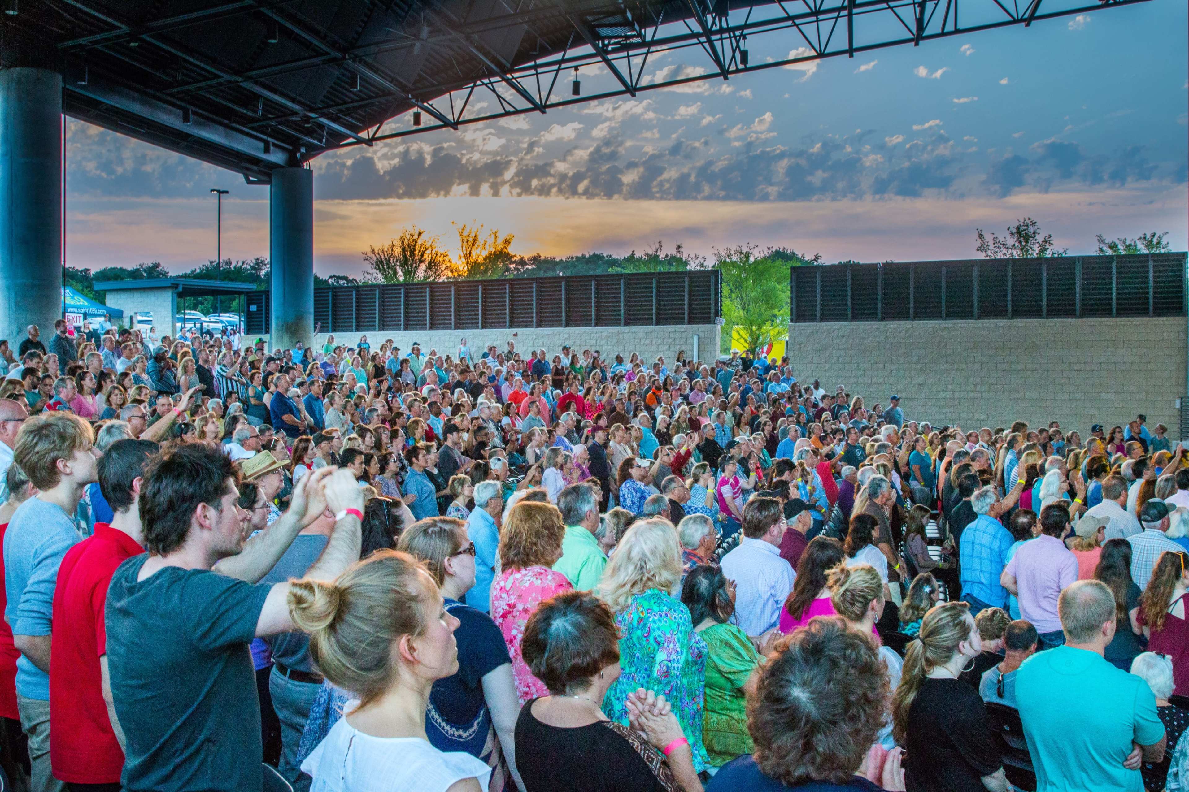 A large crowd gathers at an outdoor amphitheater during sunset, with many people clapping and enjoying a performance.