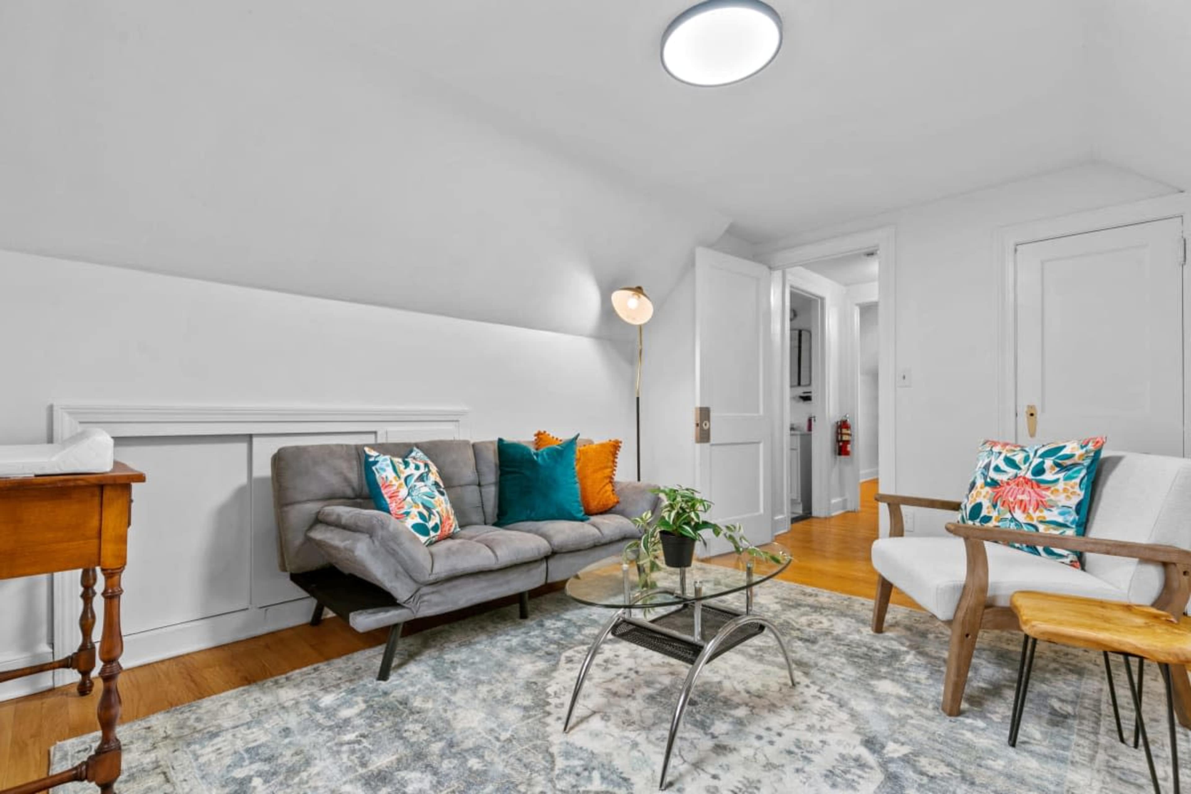 A cozy sitting area features a gray sofa with colorful cushions, a glass coffee table, and wooden accents on a textured rug.