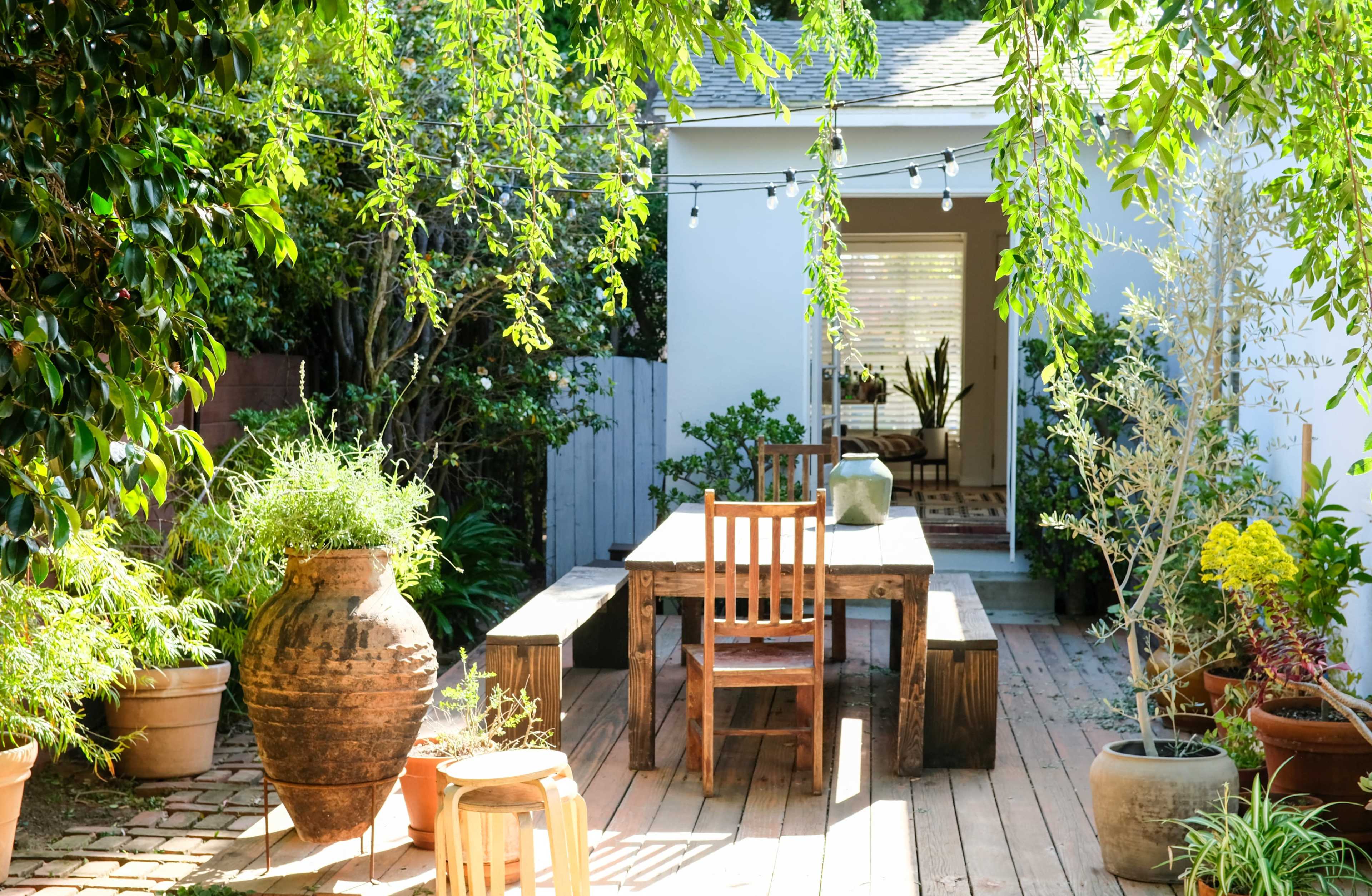 A wooden table with chairs sits on a deck surrounded by potted plants and a large ceramic pot, adjacent to a white-walled room with a window.