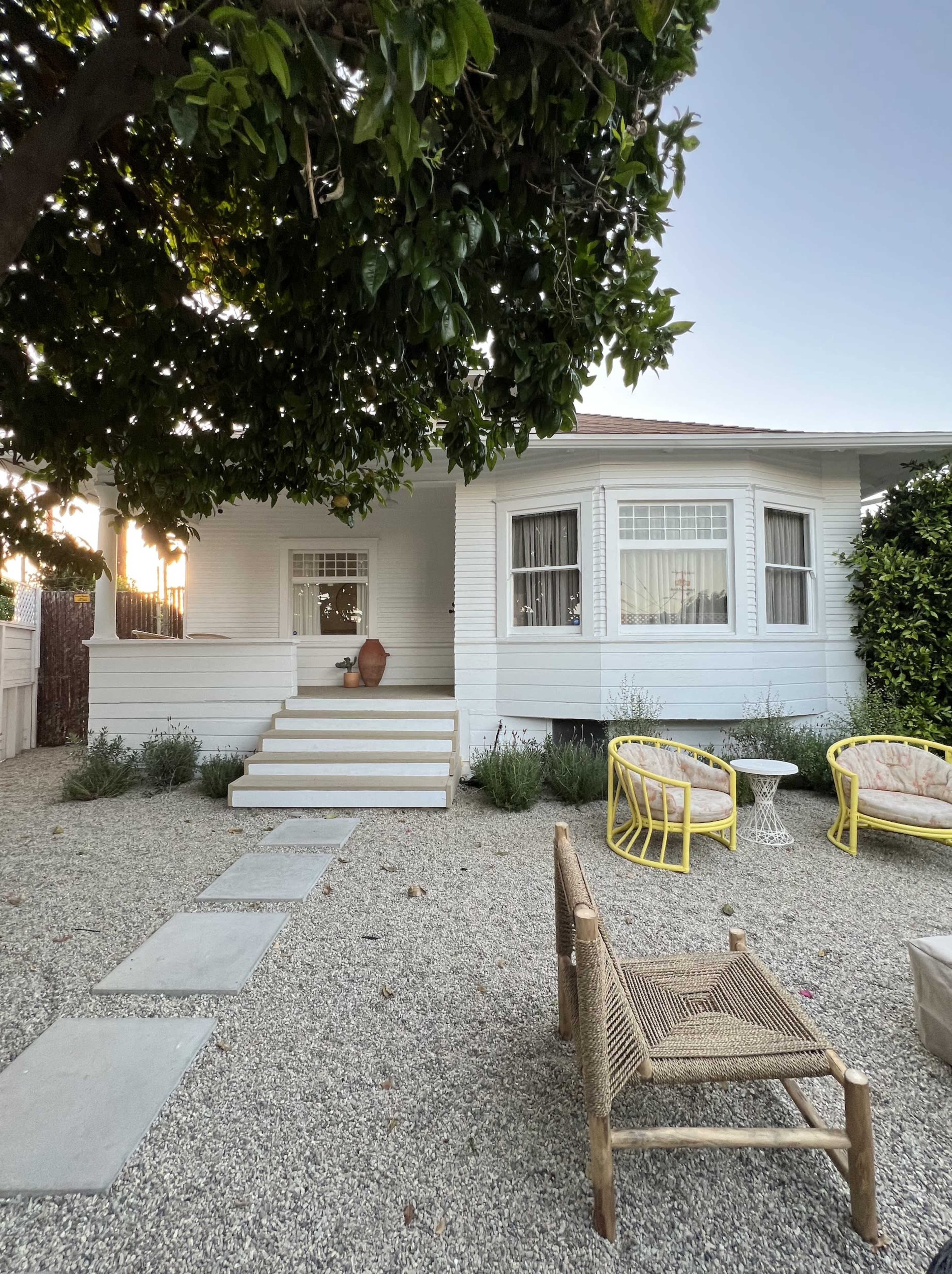 A white wooden house with a front porch is surrounded by gravel, featuring stone pathway steps and yellow outdoor furniture.