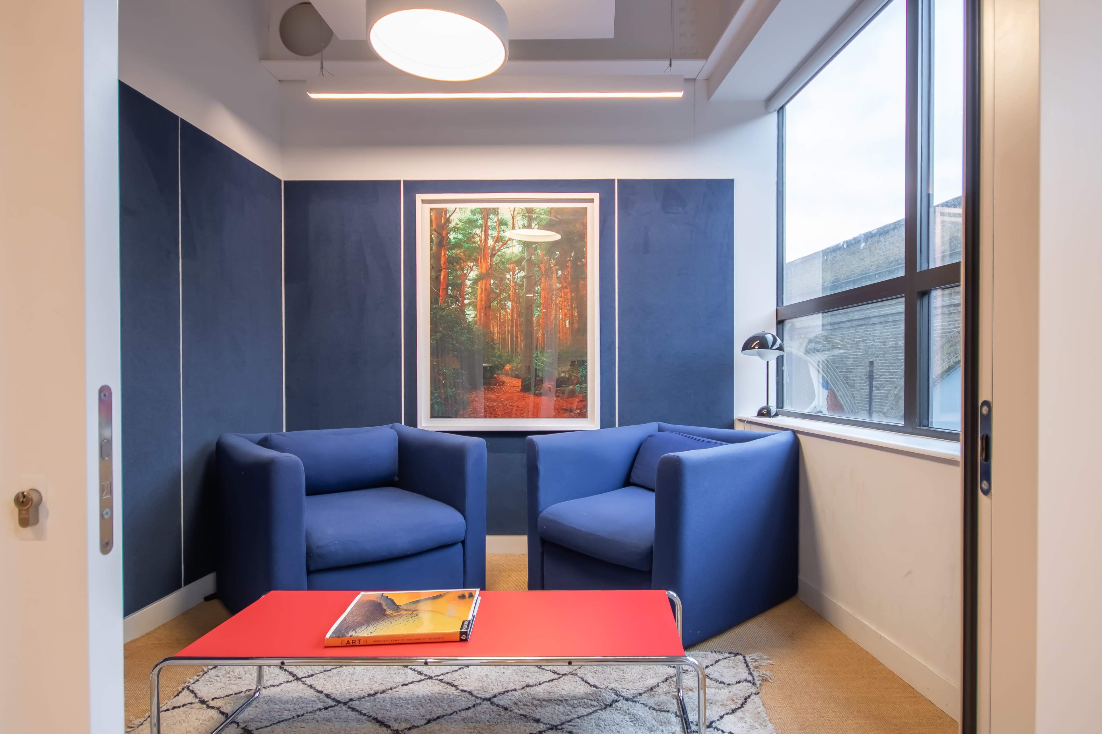 The image shows a modern seating area with two blue armchairs, a red coffee table, and a large artwork depicting a forest on the wall.