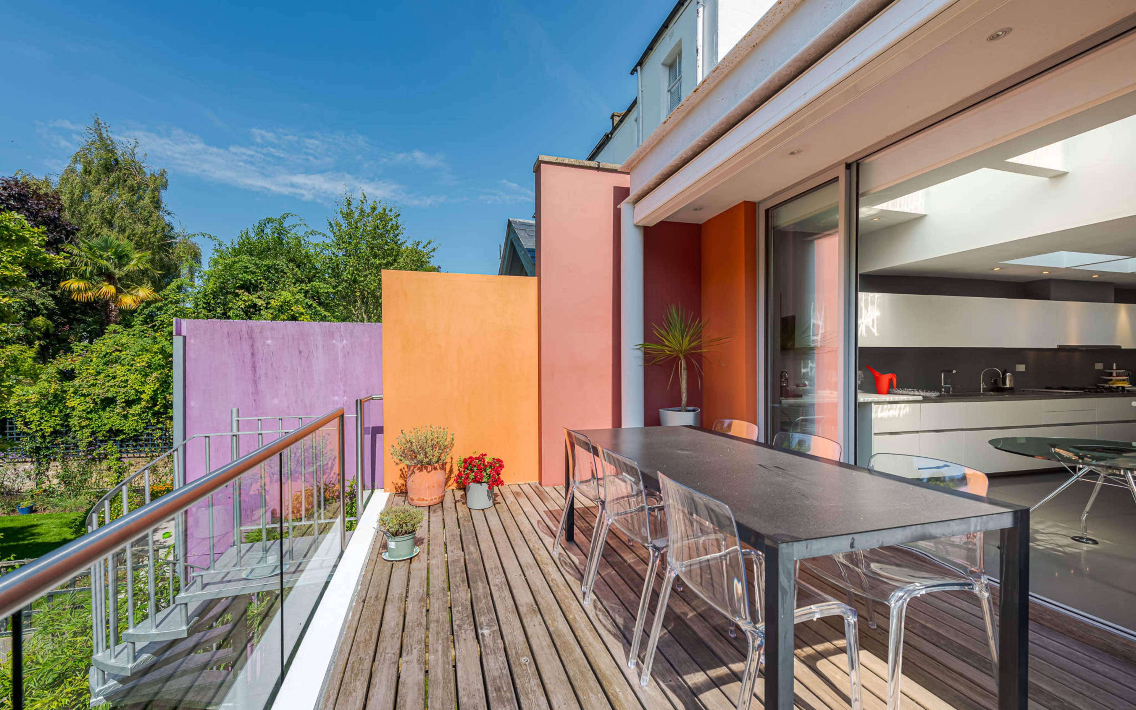 A modern deck features a large table surrounded by transparent chairs, with colorful walls and greenery in the background.