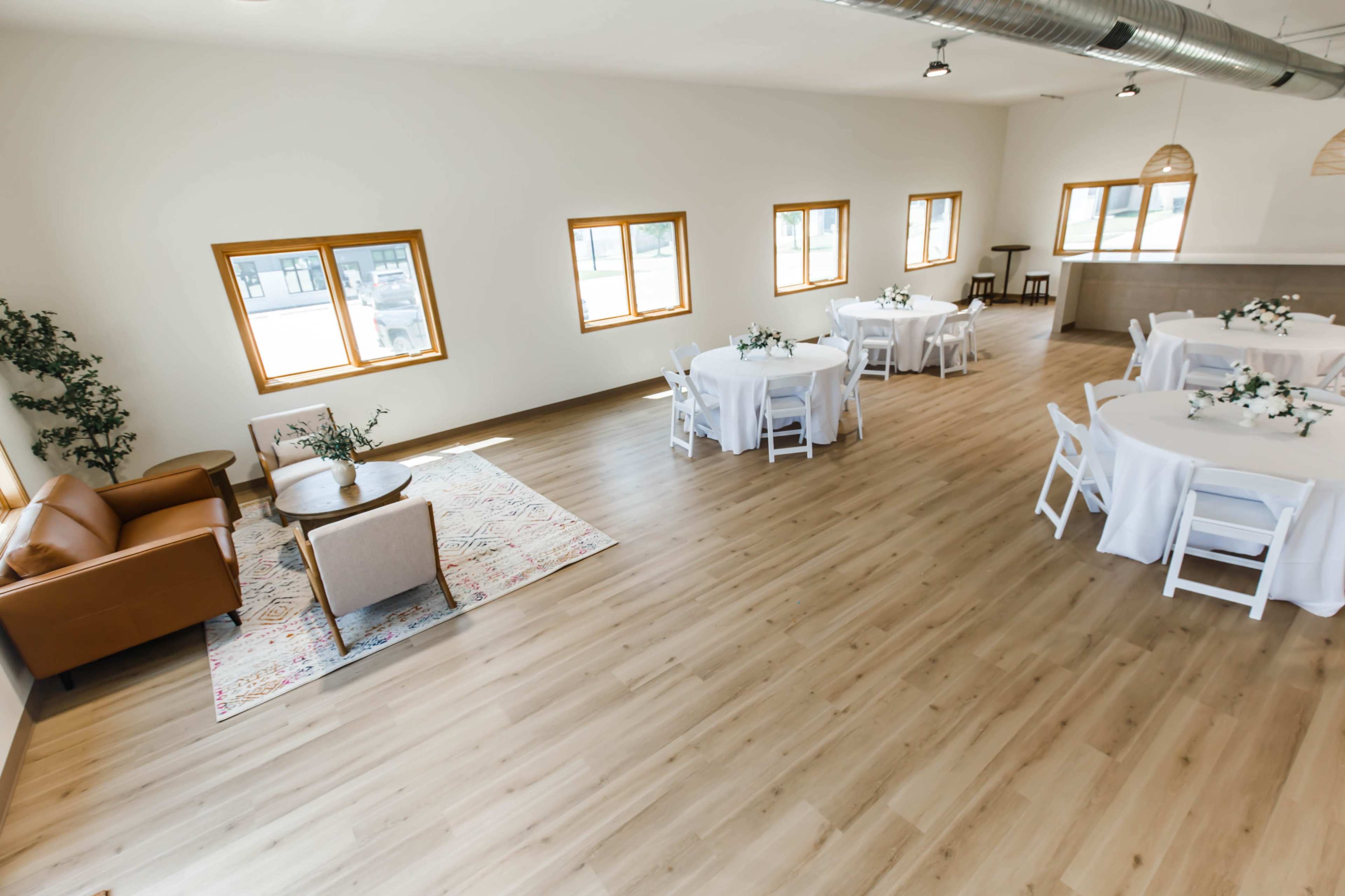 A spacious room with light-colored wooden flooring, furnished with round tables set with white tablecloths and a seating area featuring a sofa and a coffee table.