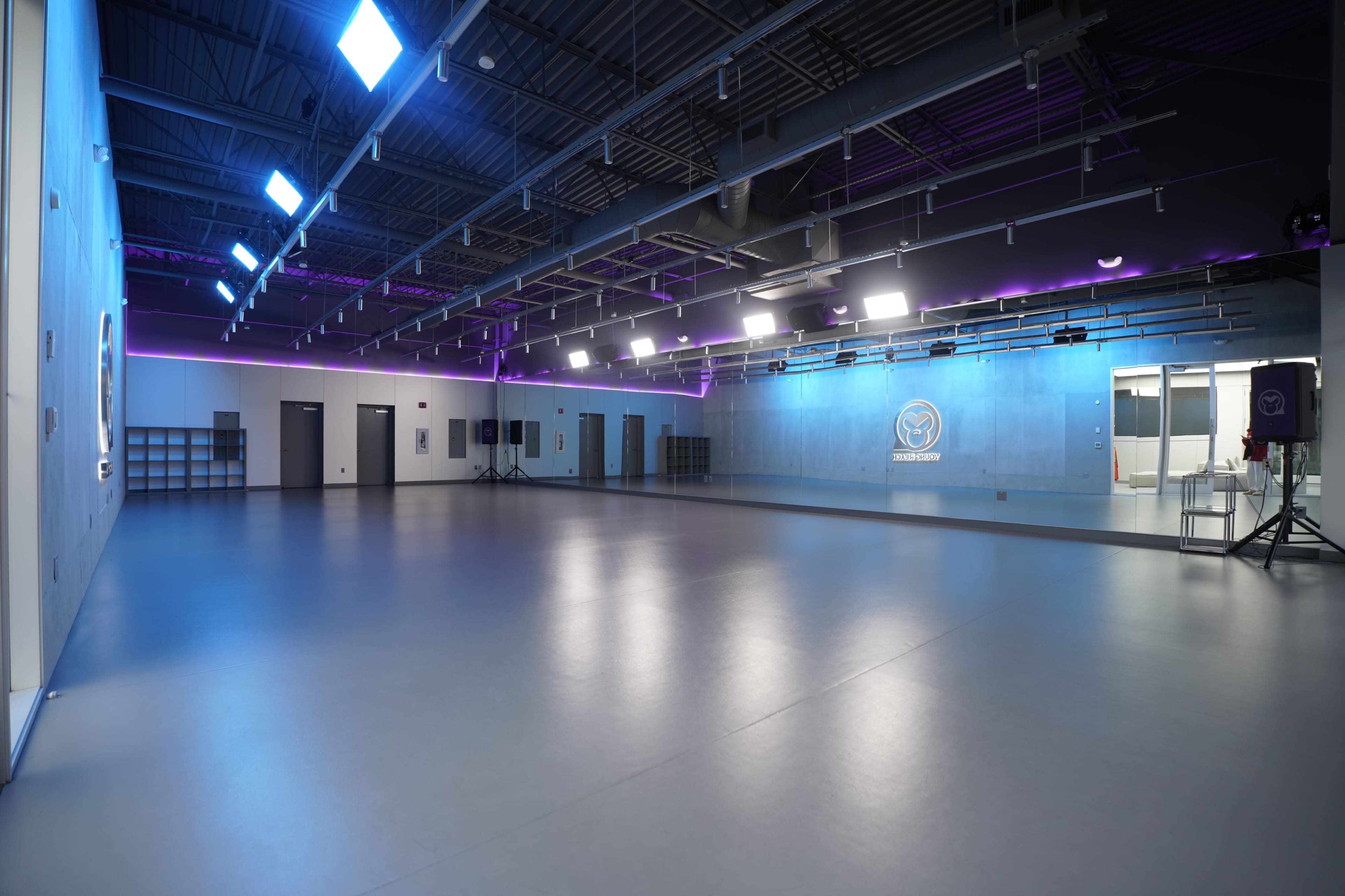 The image shows a spacious, modern dance studio with a smooth gray floor, large mirrors, and varied lighting along the ceiling.