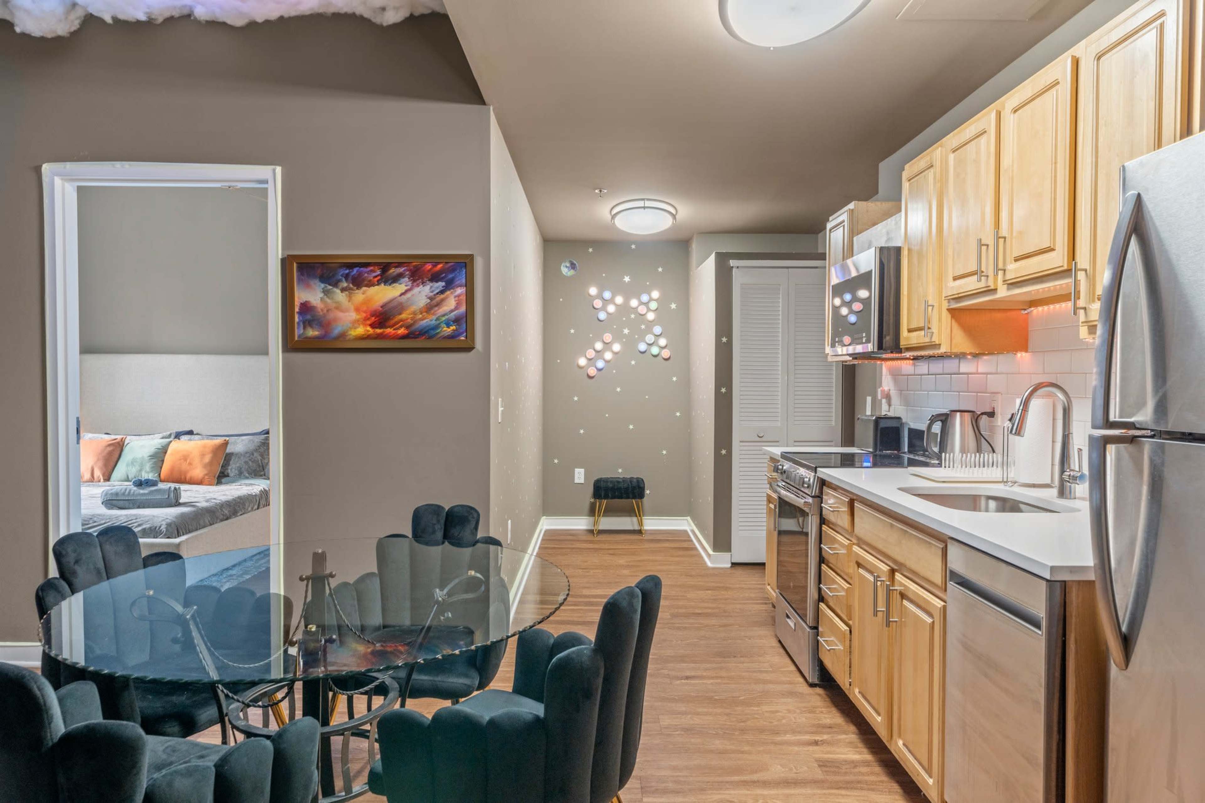 A modern kitchen area with wooden cabinets, stainless steel appliances, and a round glass dining table surrounded by dark upholstered chairs.