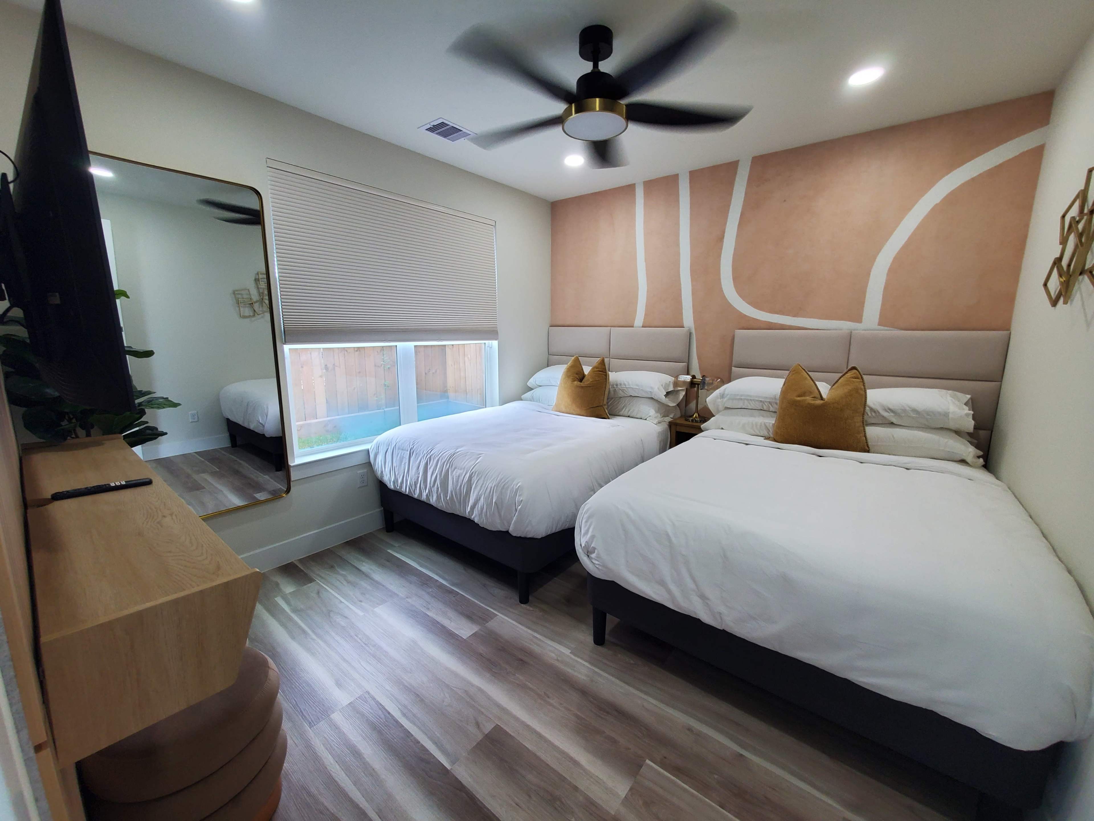 The image shows a stylish bedroom with two queen-sized beds, a large mirror, and contemporary decor featuring a warm wall design.