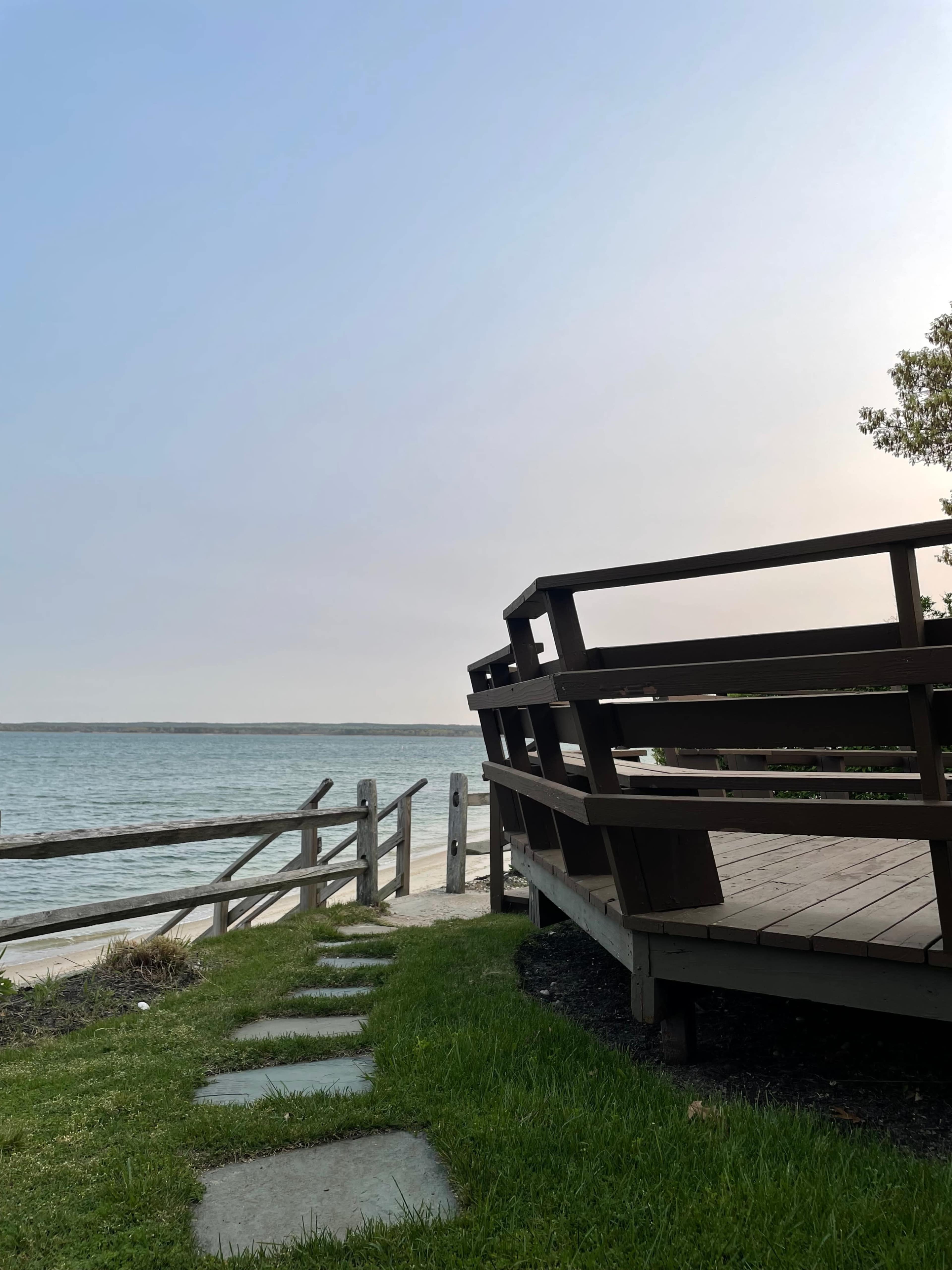 A wooden deck overlooks a calm body of water, with stepping stones leading through grassy areas to a shoreline.