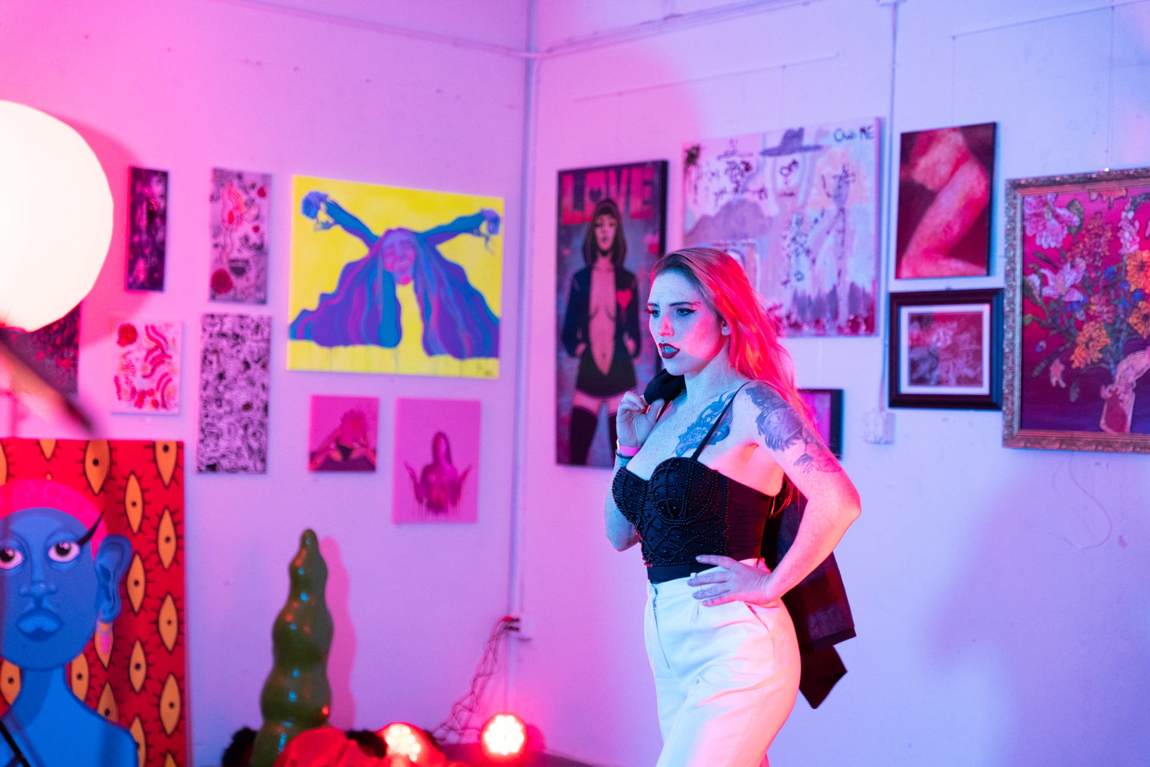 A model poses in front of a gallery wall filled with colorful artwork while soft, colored lights illuminate the space.
