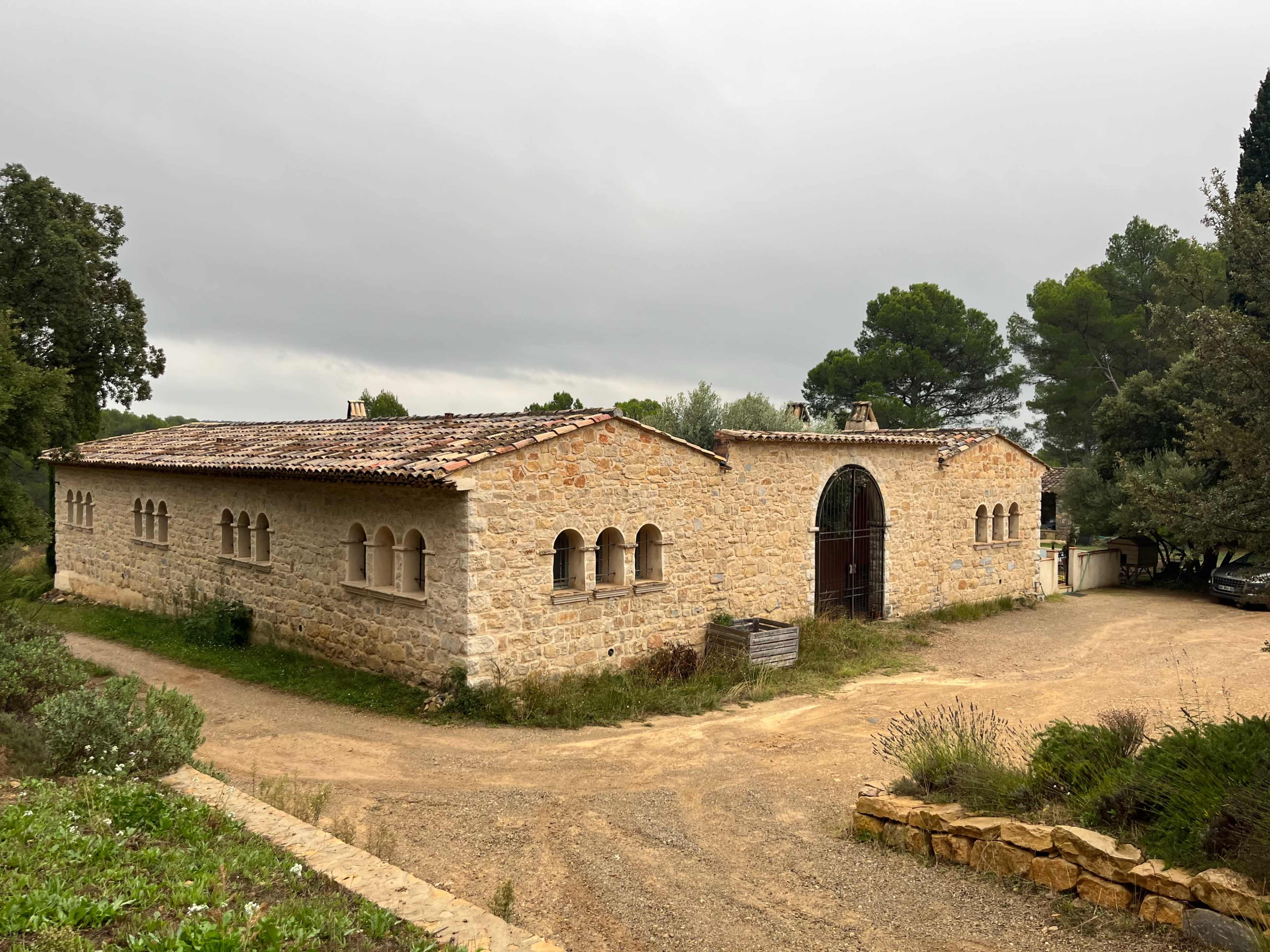 A stone building with arched windows and a large double door is set beside a gravel pathway surrounded by trees.