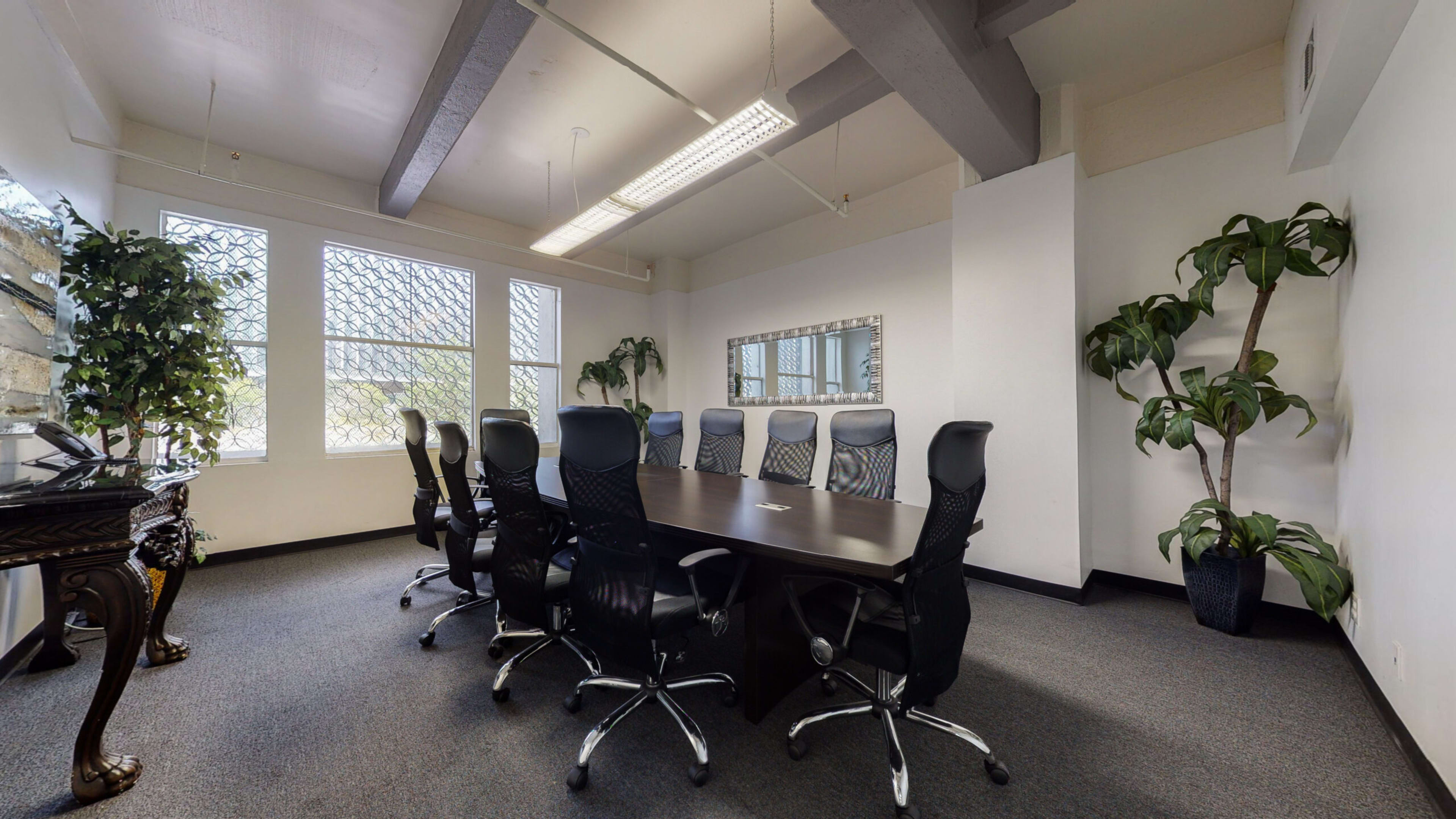 The image shows a conference room with a long table surrounded by black office chairs, large windows, and potted plants.