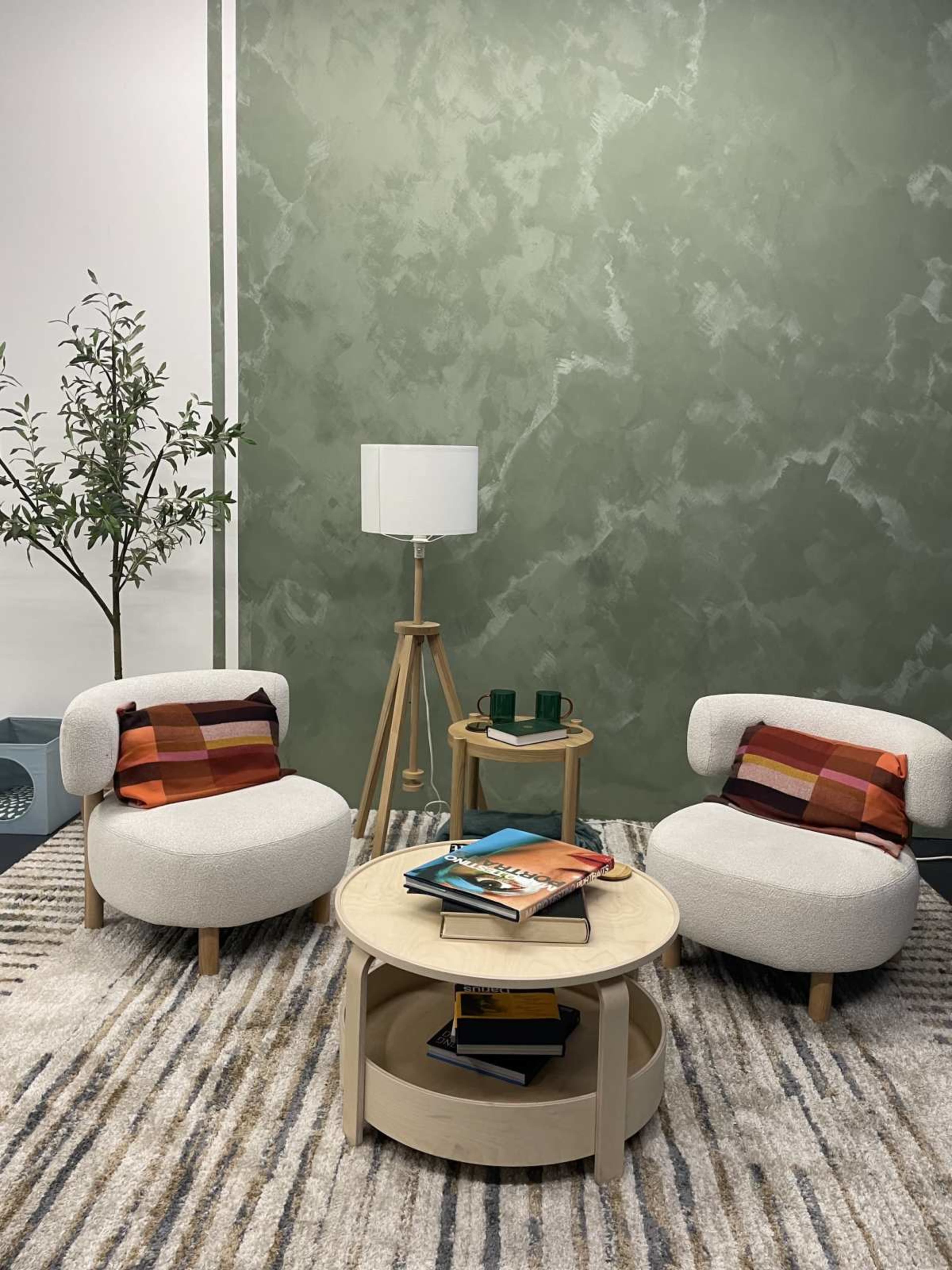 The image features a cozy seating area with two upholstered chairs, a round coffee table stacked with magazines, a floor lamp, and a textured green wall in the background.