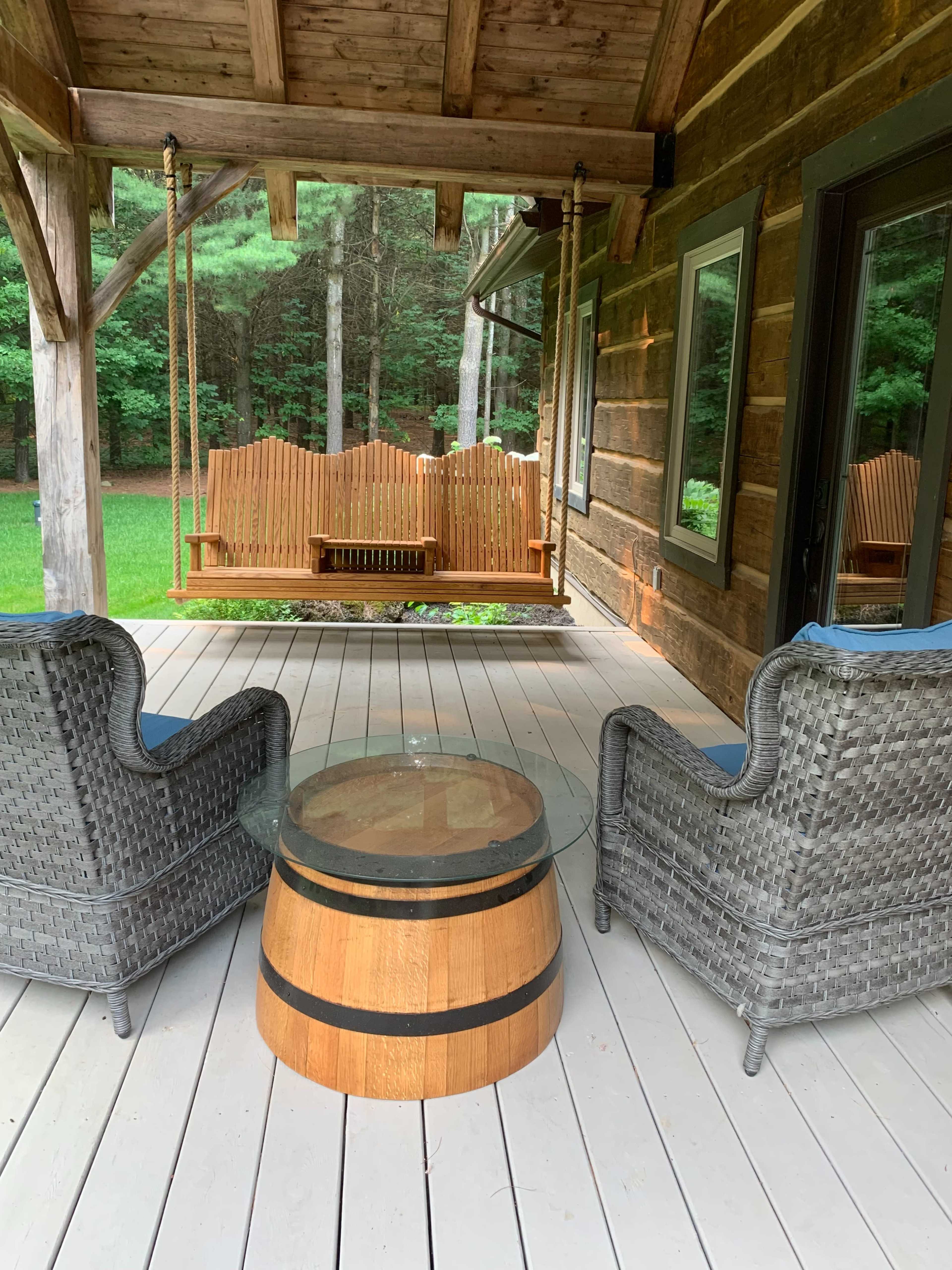 A porch features two wicker chairs and a wooden barrel coffee table with a glass top, overlooking a swing and a green yard.