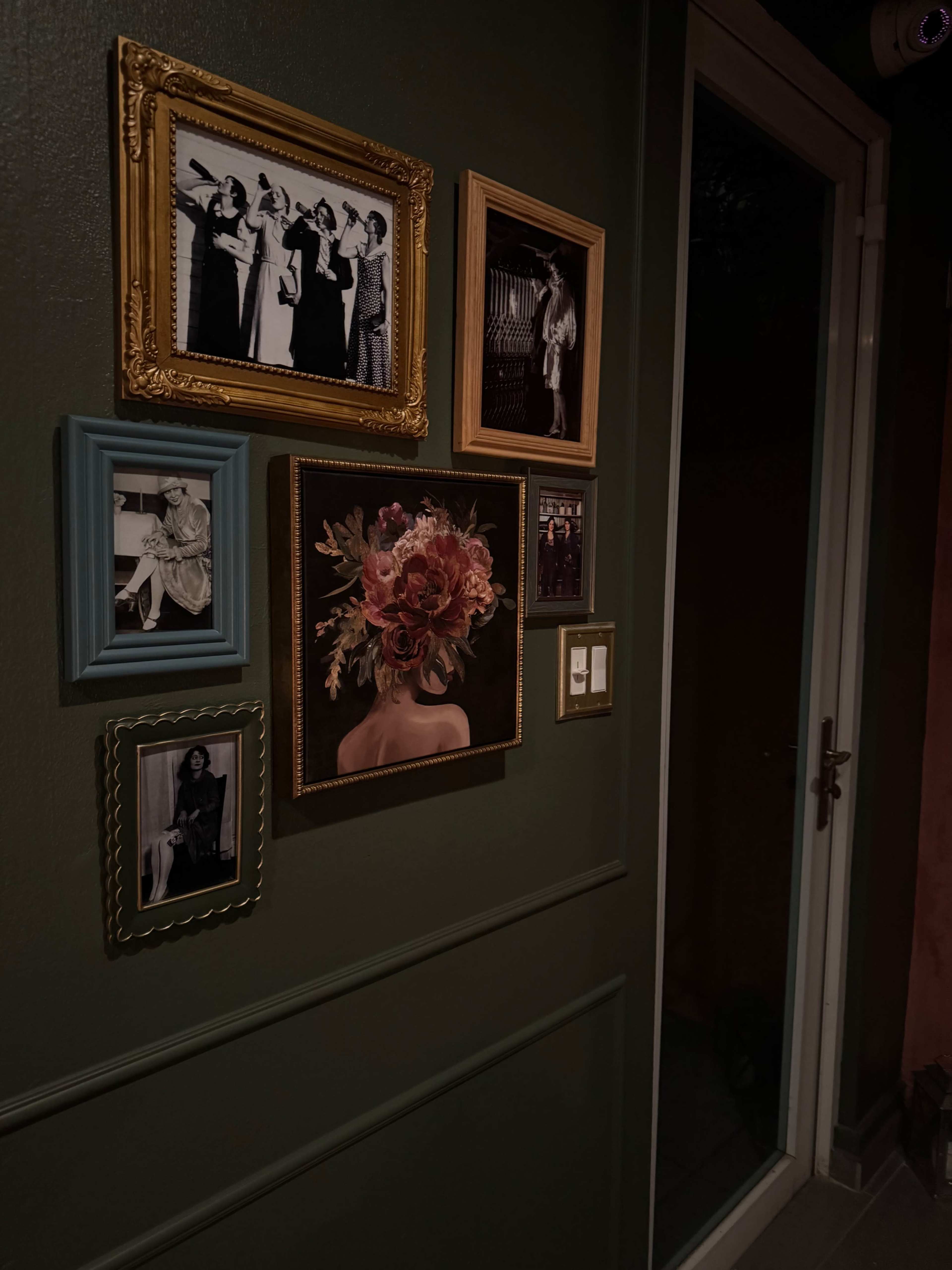 The image shows a collection of framed photographs and artwork arranged on a dark green wall near a doorway.