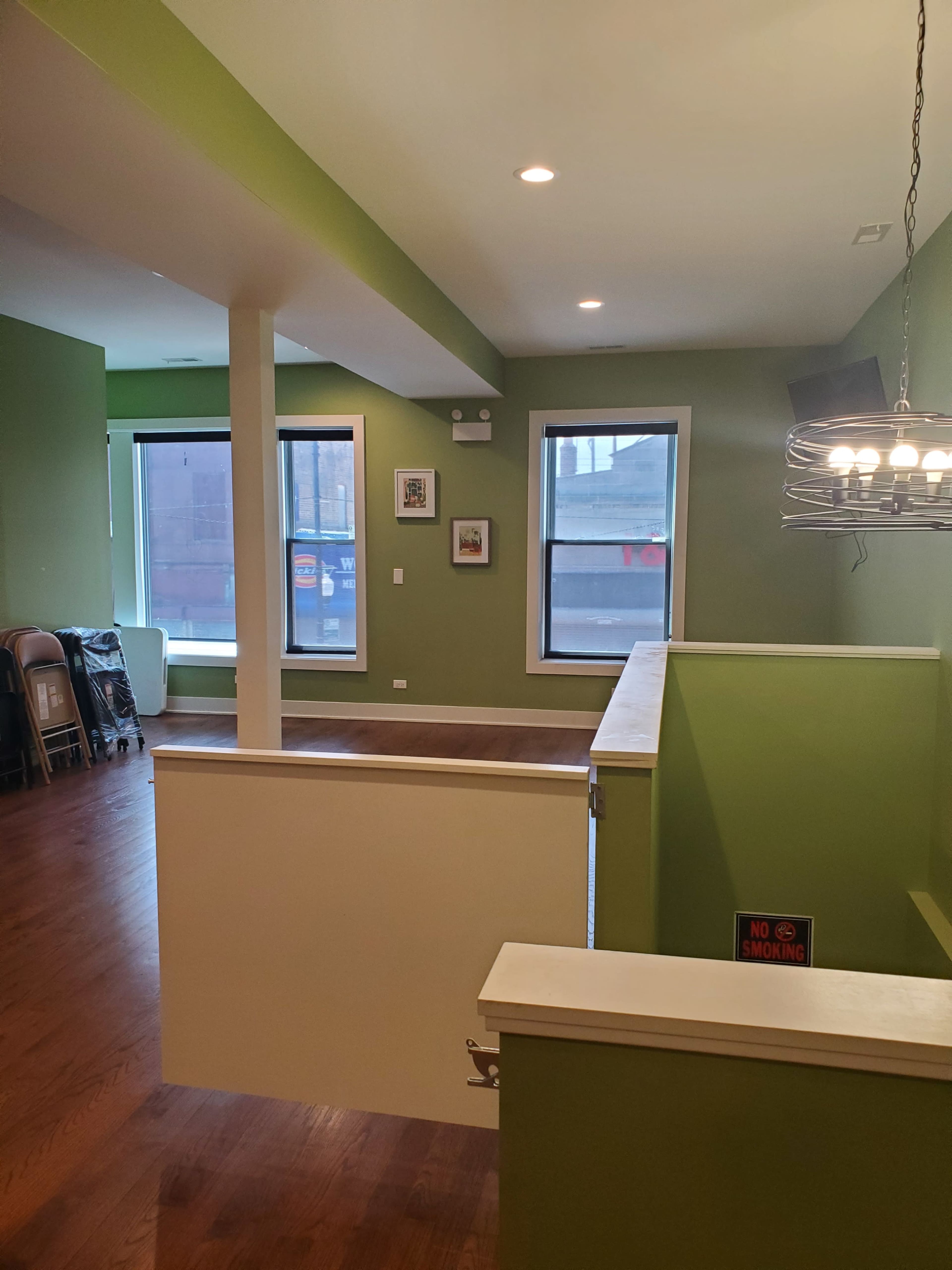 The image shows a spacious, green-walled room with wooden flooring, featuring large windows and a seating area with chairs in the corner.