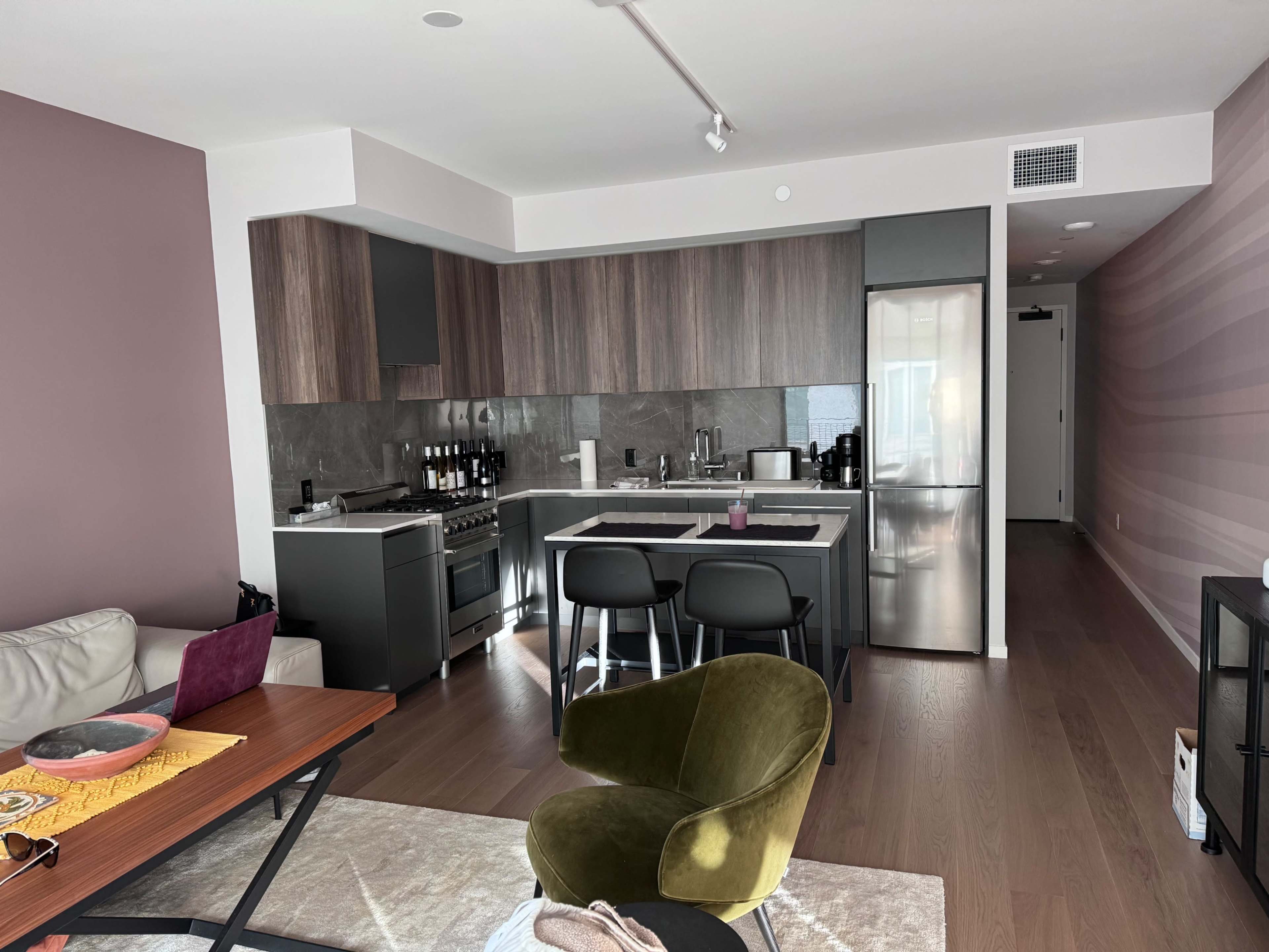 A modern kitchen with dark wood cabinets, stainless steel appliances, and a light-colored living area featuring a coffee table and a green chair.