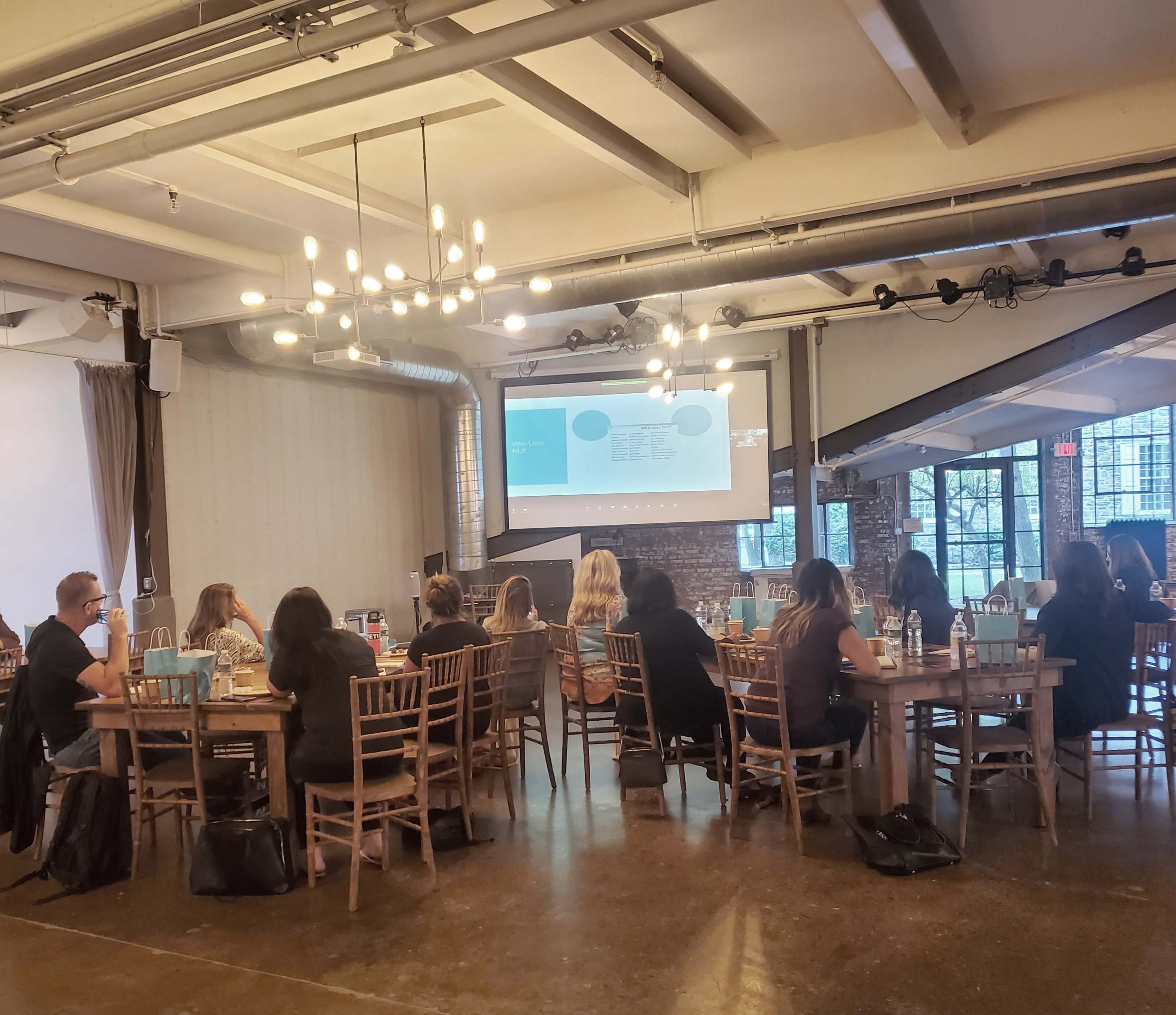 A group of people is seated at tables in a well-lit room, attentively listening to a presentation displayed on a screen.