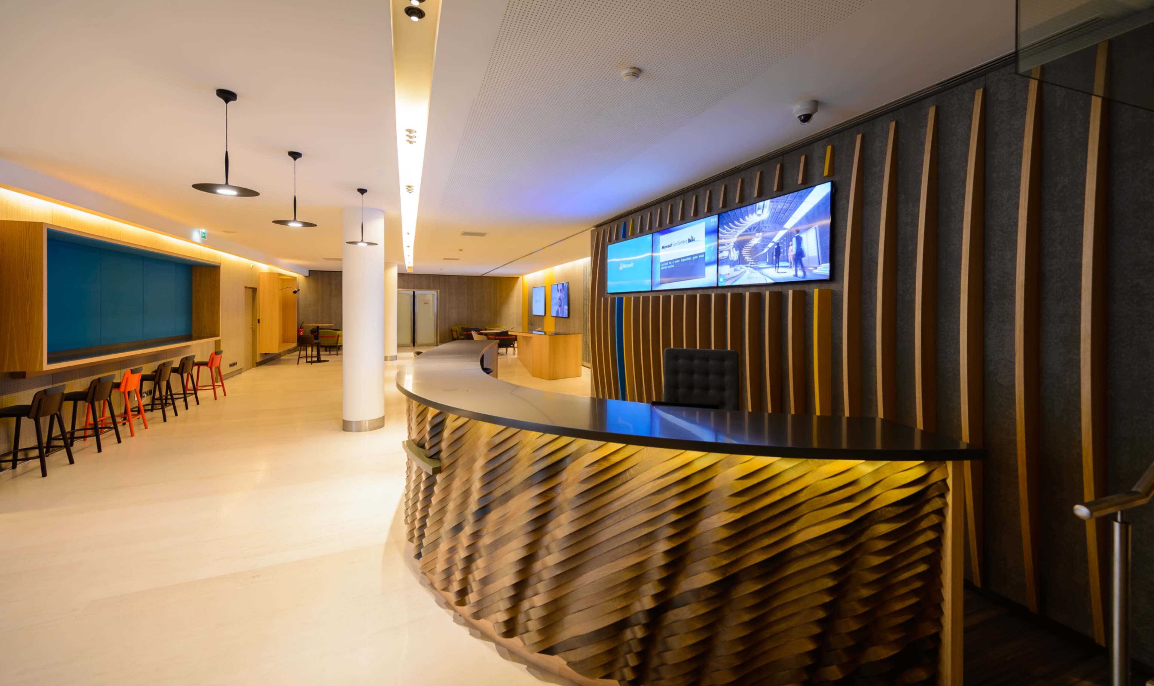 The image shows a modern reception area with a sculptural front desk, wooden paneling, and digital screens displaying images.
