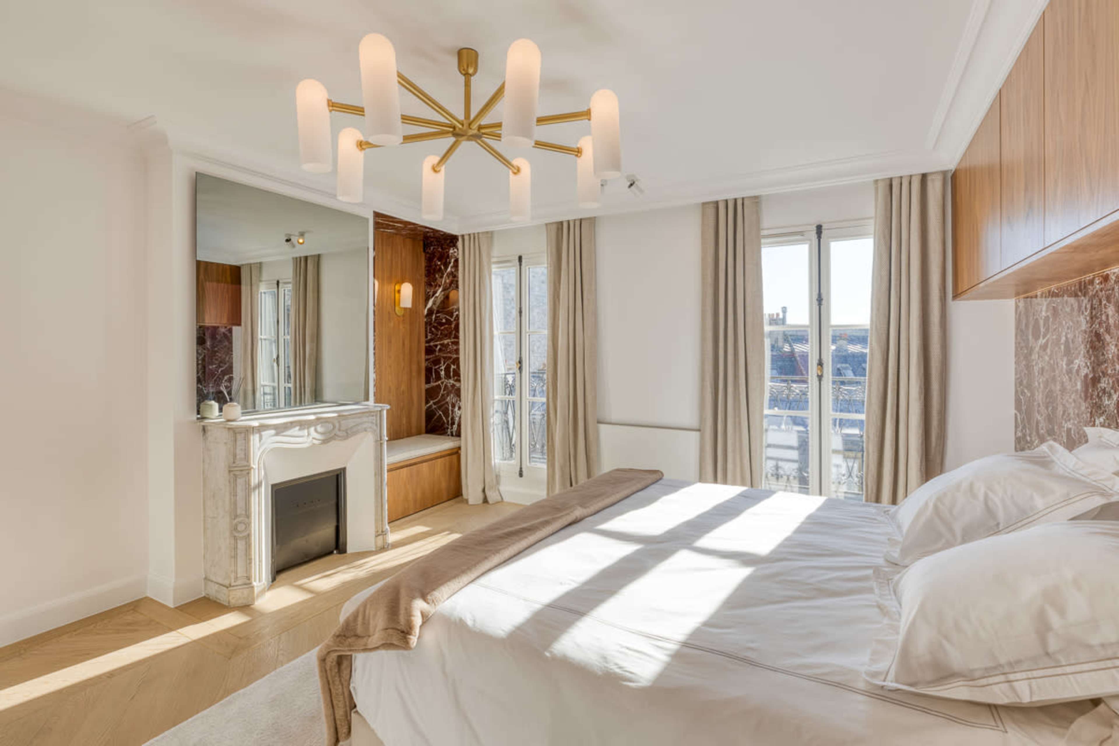 The image shows a modern bedroom featuring a bed, a marble fireplace, large windows with curtains, and a stylish overhead light fixture.