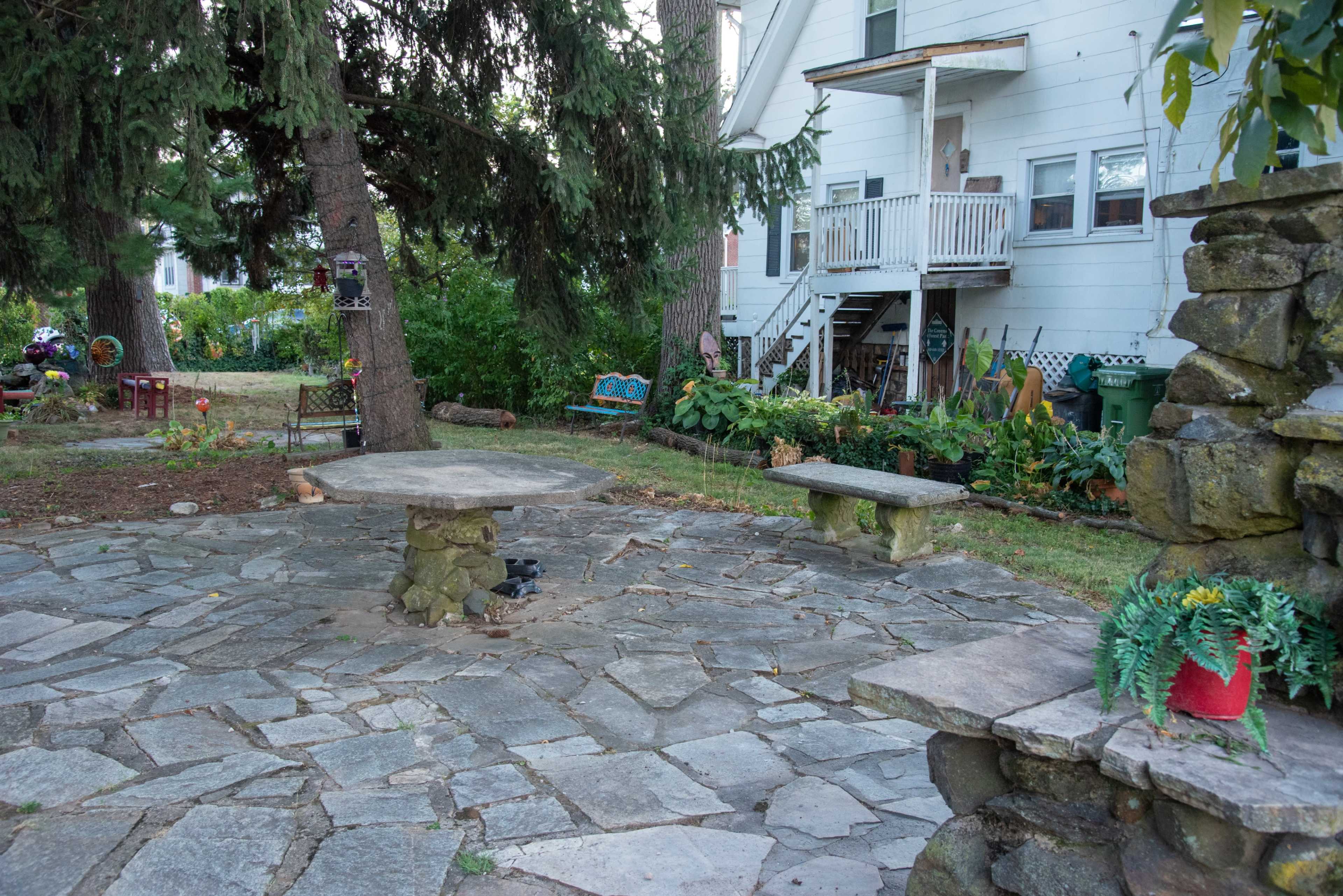 The image shows a stone patio with a circular table and benches surrounded by greenery and a house in the background.