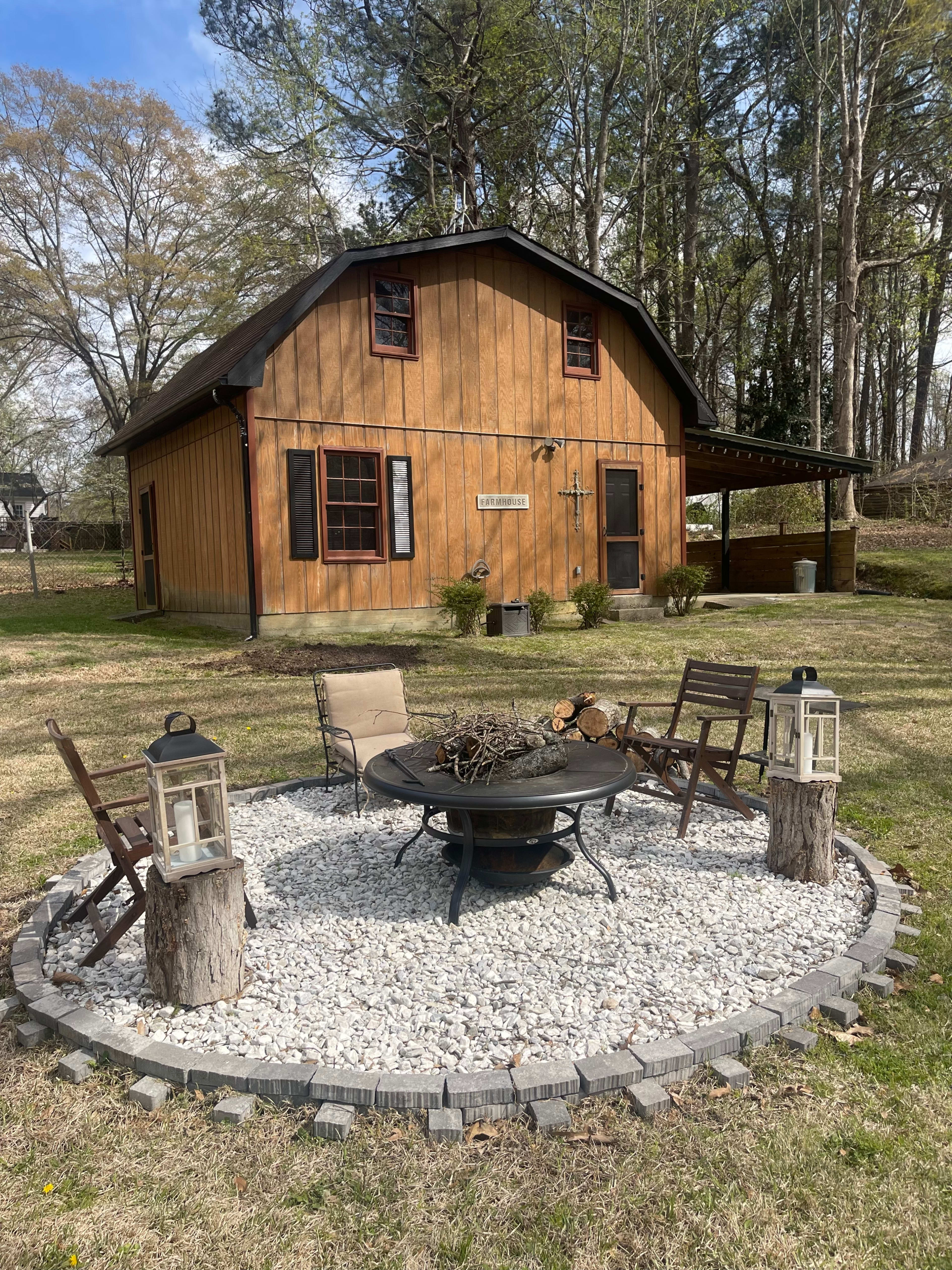 A fire pit surrounded by four chairs and lanterns is situated in a gravel circle in front of a two-story wooden building.